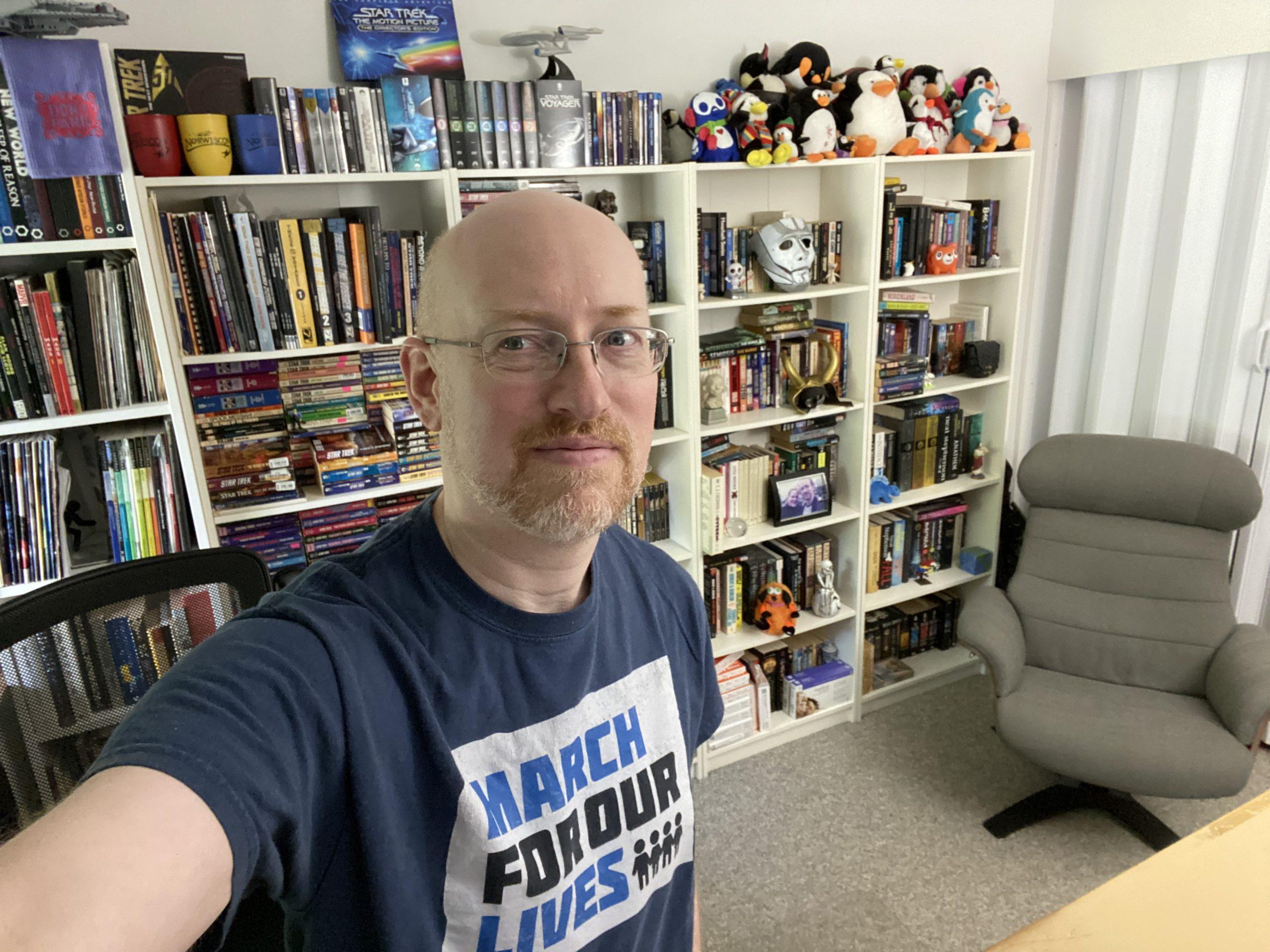 Me standing in my office. The wall behind me is lined with white bookcases filled with books, plus various toys and knicknacks. The top shelf has Star Trek DVDs on one half, lots of stuffed penguins on the other half. In front of a closet is a grey chair that somewhat resembles the chairs on the bridge of the Enterprise 1701-D.