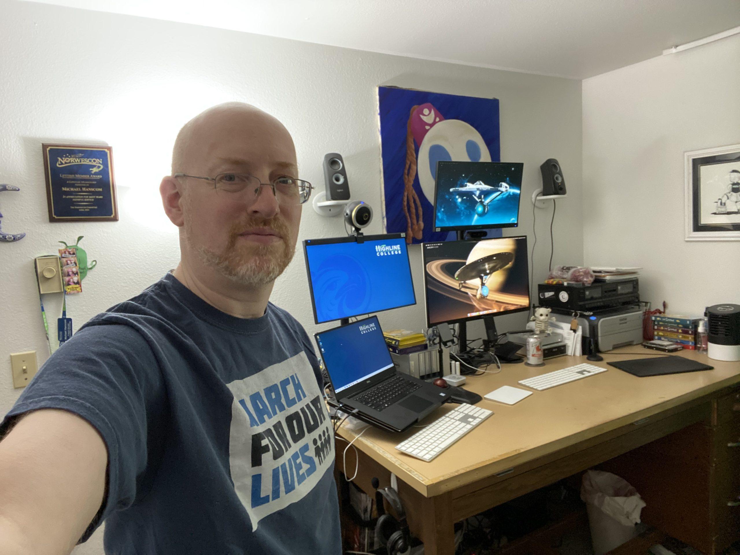 Me standing in front of my desk, a repurposed large drafting table. The left third of the desk holds my work laptop under a second monitor, the middle third holds two monitors mounted one above the other, the right third holds a printer under a dual audio cassette deck. Comptuer speakers are mounted on the wall on either side of a large painting of a round white face wearing a fez. Also on the wall is a cartoon sketch of me and a plaque from Norwescon.