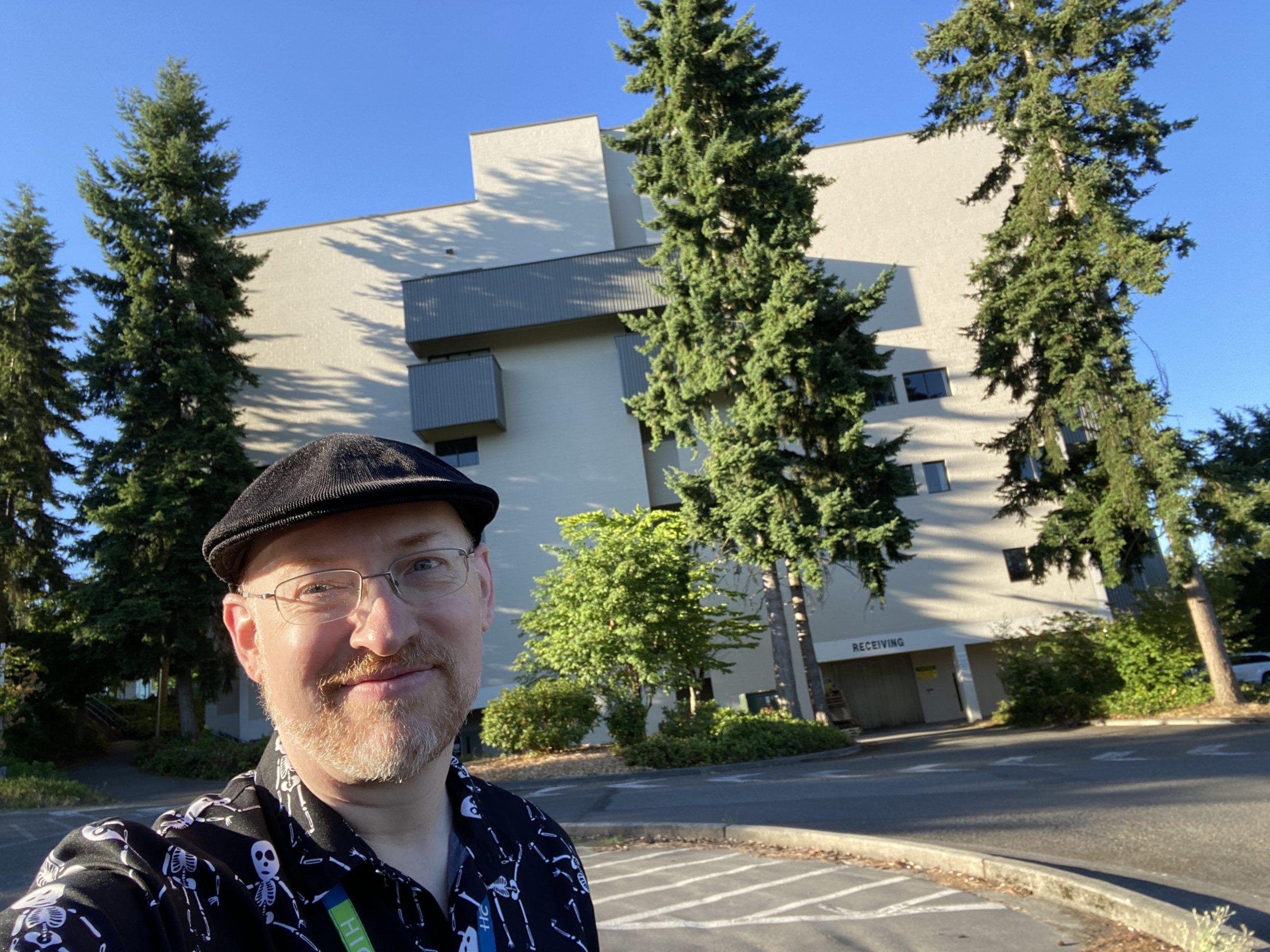 Me standing in a parking lot outside the Highline College library, a very square, somewhat brutalesque 1960s-era building, on a sunny morning.