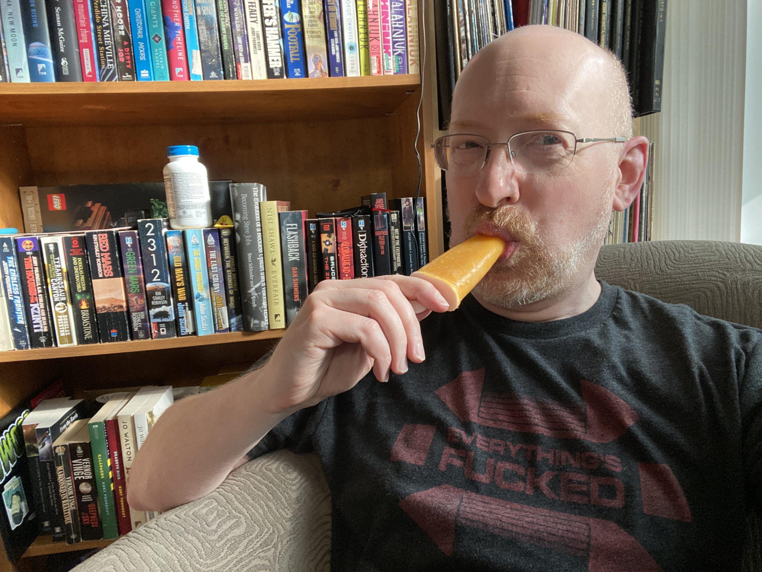 Me sitting in a green chair in front of a bookshelf sucking on an orange popsicle.