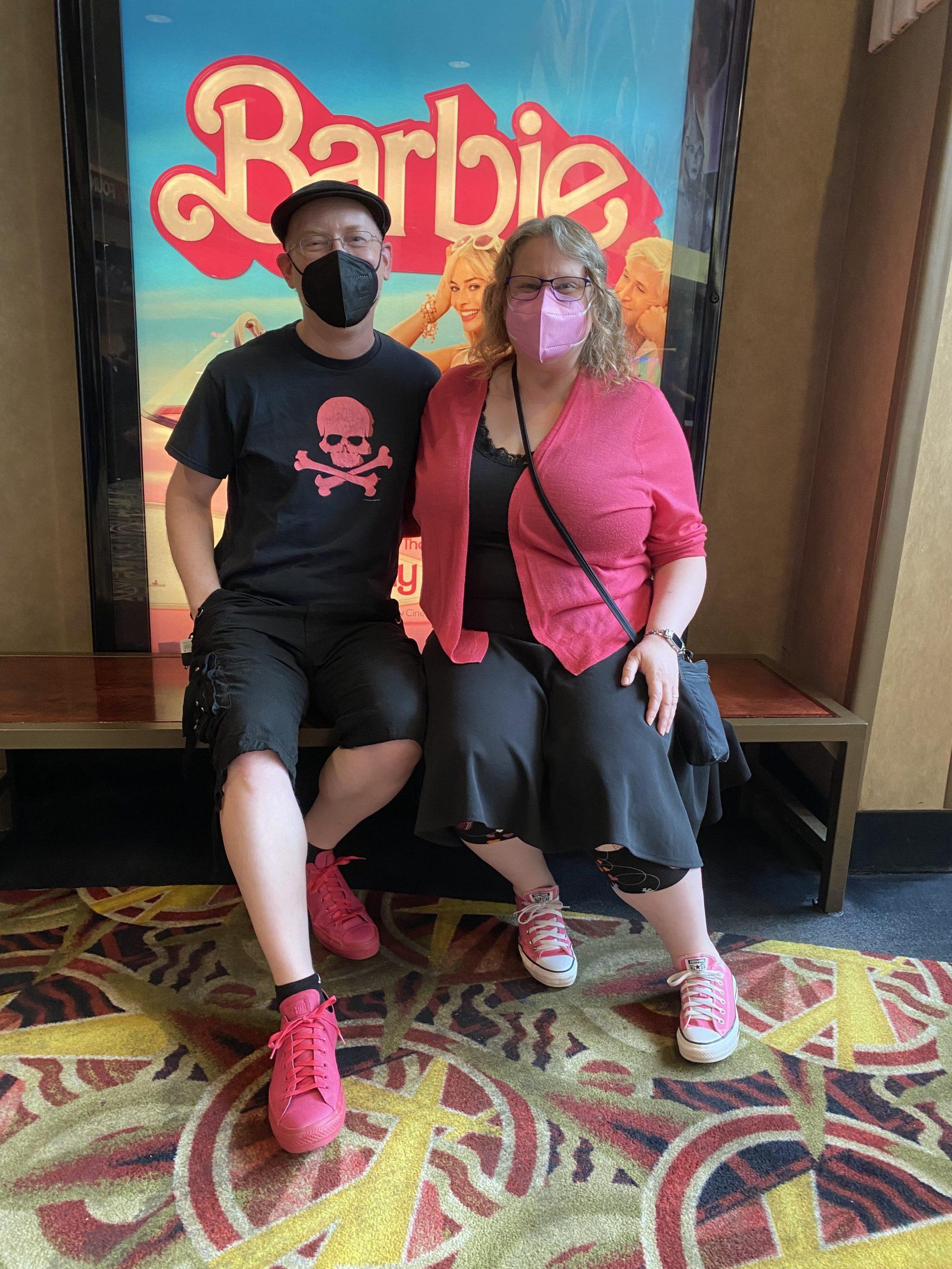 My wife and I sitting in front of a poster for the Barbie movie. I'm wearing black shorts and a black t-shirt with a pink skull and crossbones and bright pink Converse, my wife is wearing a pink sweater over a black top and skirt and light pink Converse.