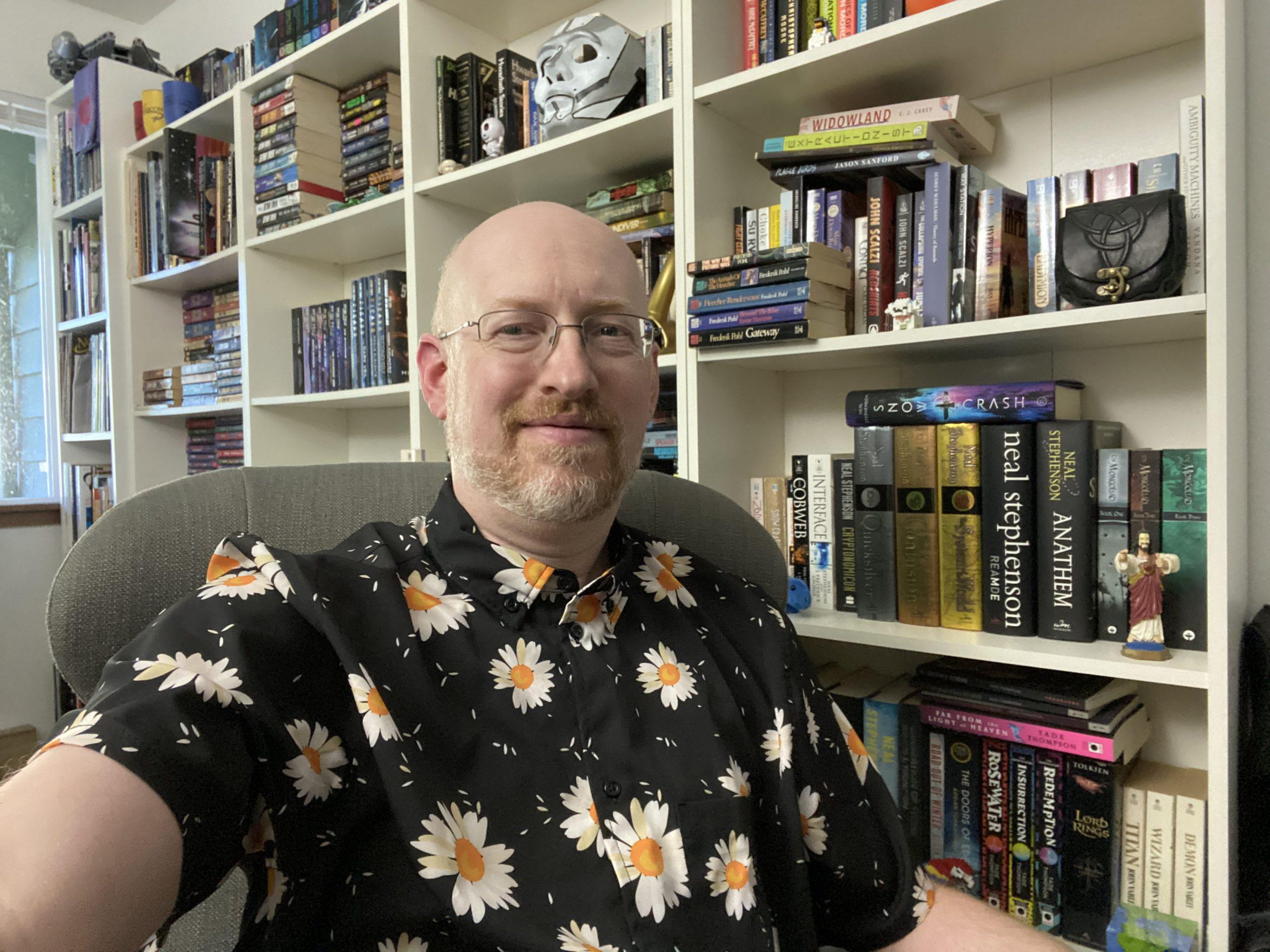 Me wearing a black shirt with daisies printed on it, sitting in a grey chair in front of several white bookcases, all filled with books and knicknacks.