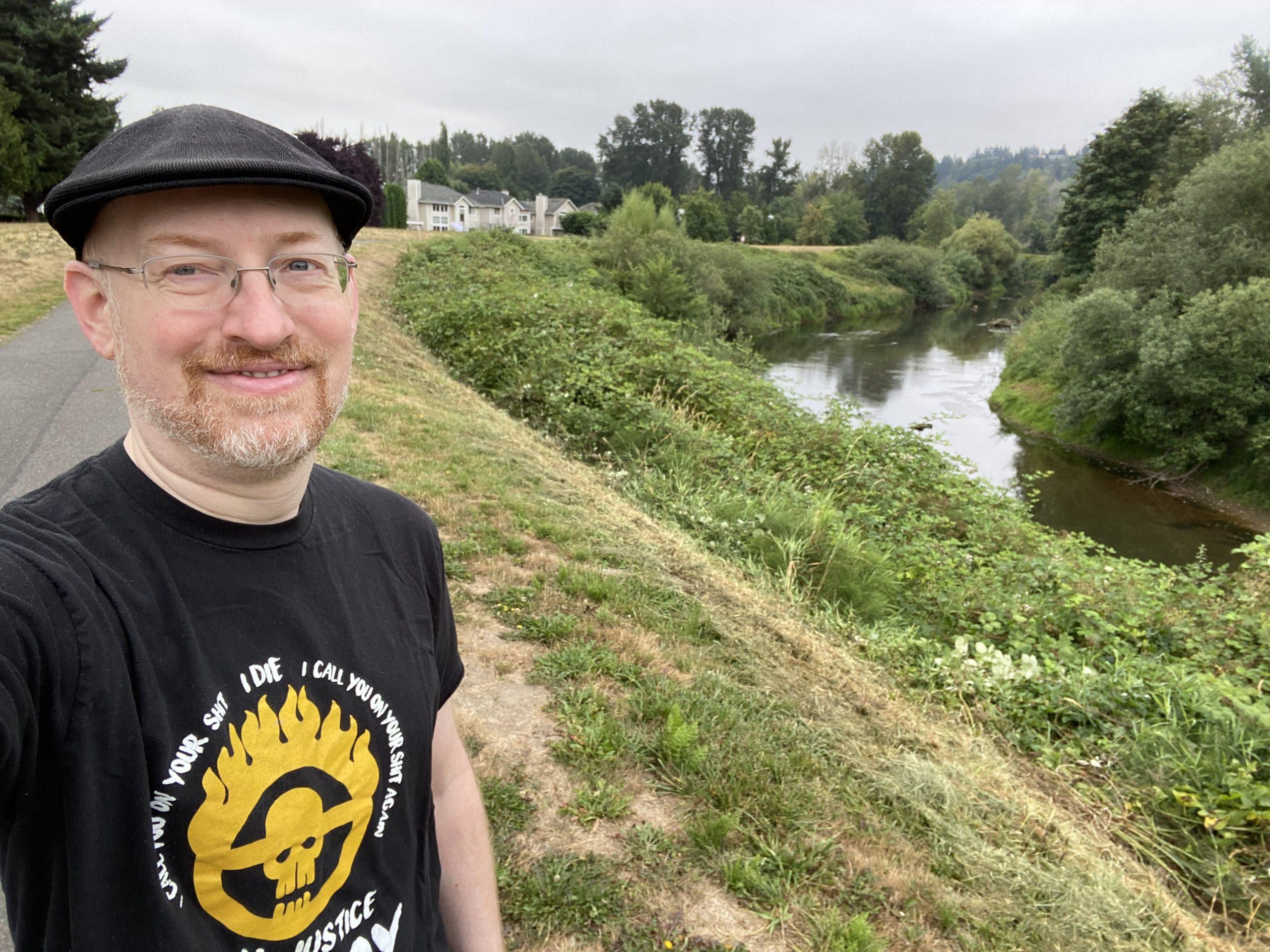 Me standing by the side of a pathway running alongside the bank of a river on a grey cloudy morning. The bank is overgrown with green plants and trees.