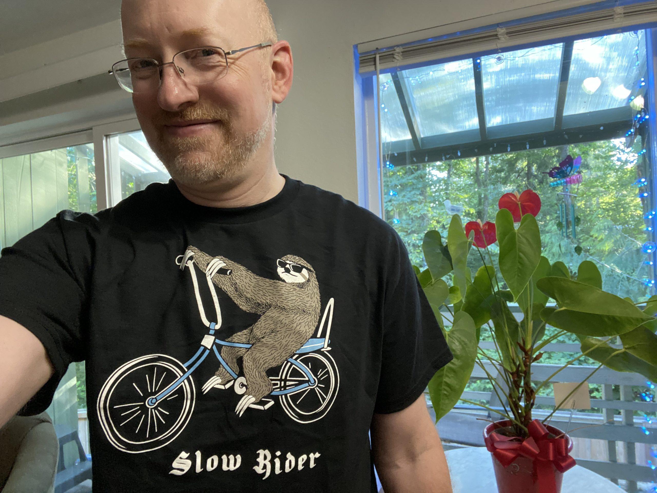 Me in our living room in front of a window, smiling and wearing a black t-shirt with a drawing of a sloth riding a bicycle and the text 'slow rider'.