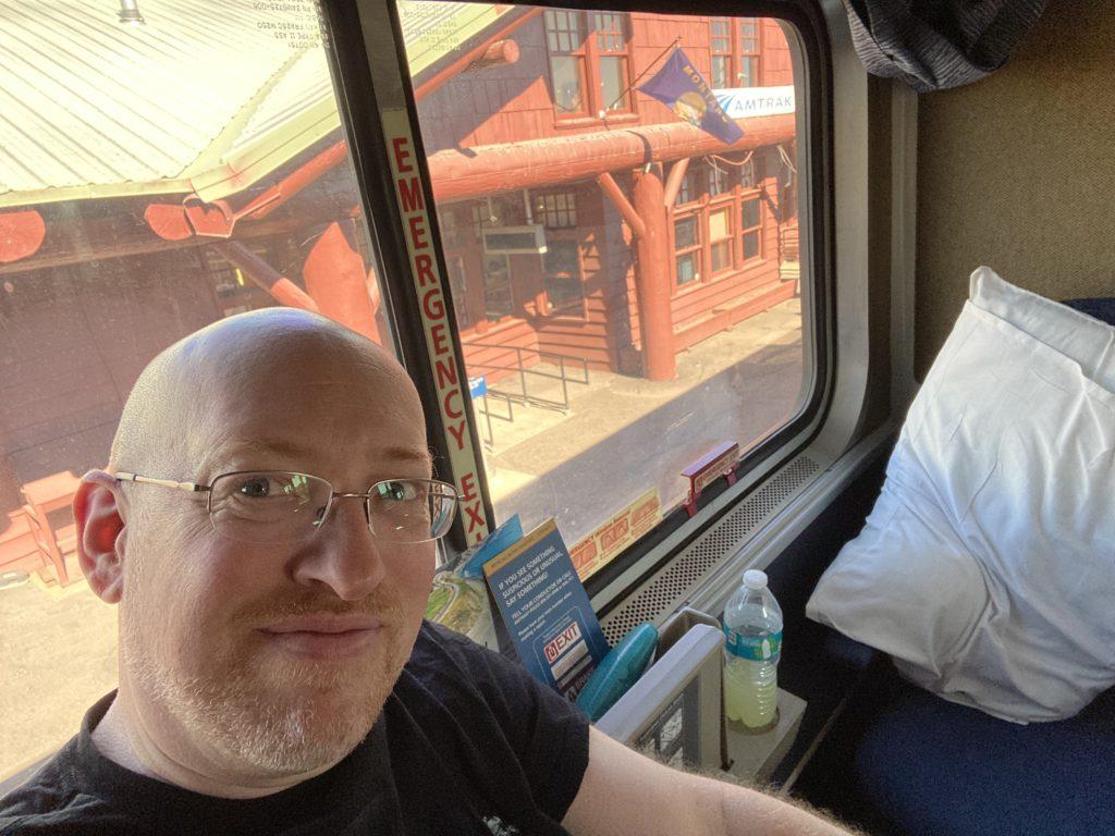 Me in a sleeper cabin on a train. An Amtrak station flying a Montana flag is visible through the window behind me.