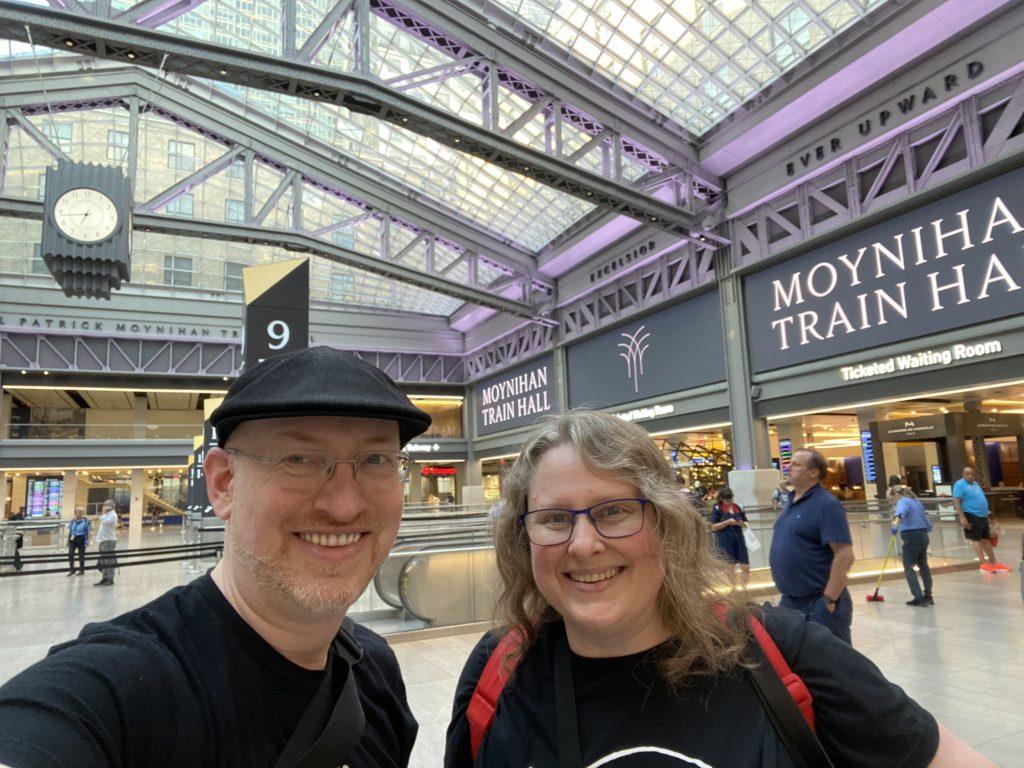 My wife and me standing in the main lobby of New York City’s Moynihan Train Hall. It is a large space with steel and glass roof.