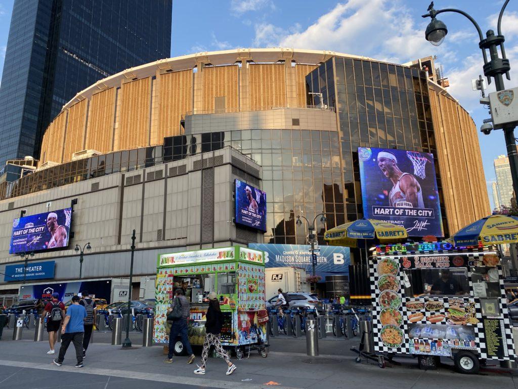 The outside of Madison Square Garden, a large, round, tan colored stadium. Two hod dog vendor stands are on the sidewalk in front as people walk by.