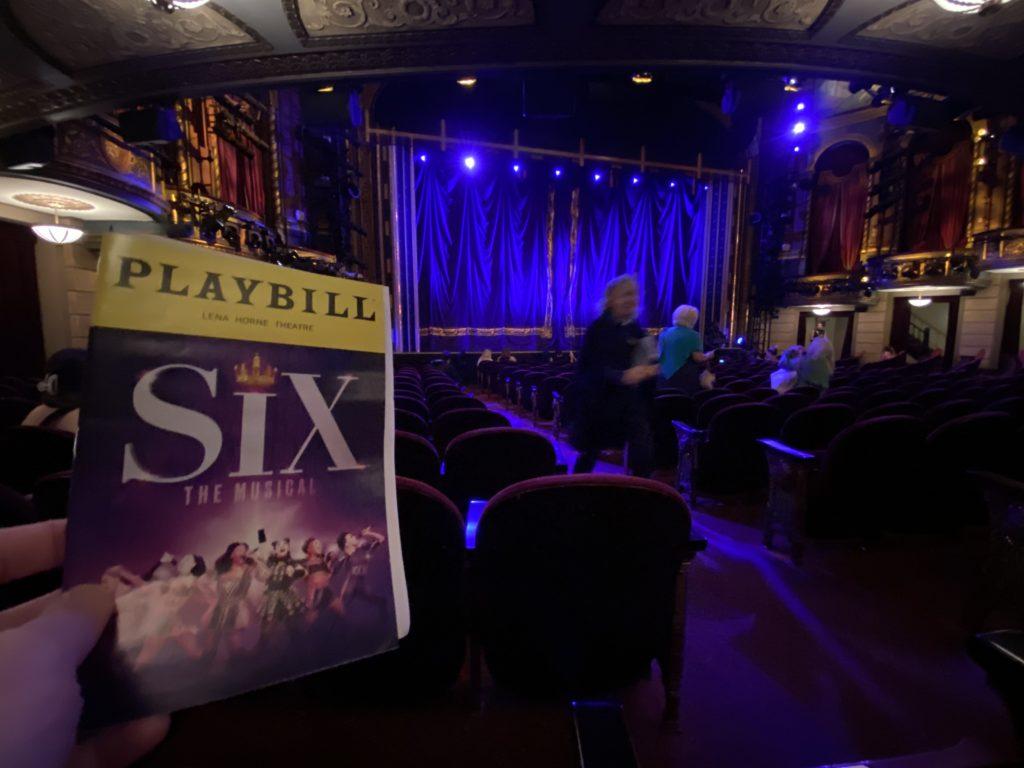 The inside of an ornate theater lit in blues and purples. The Playbill for the musical Six is being held up into the photo.