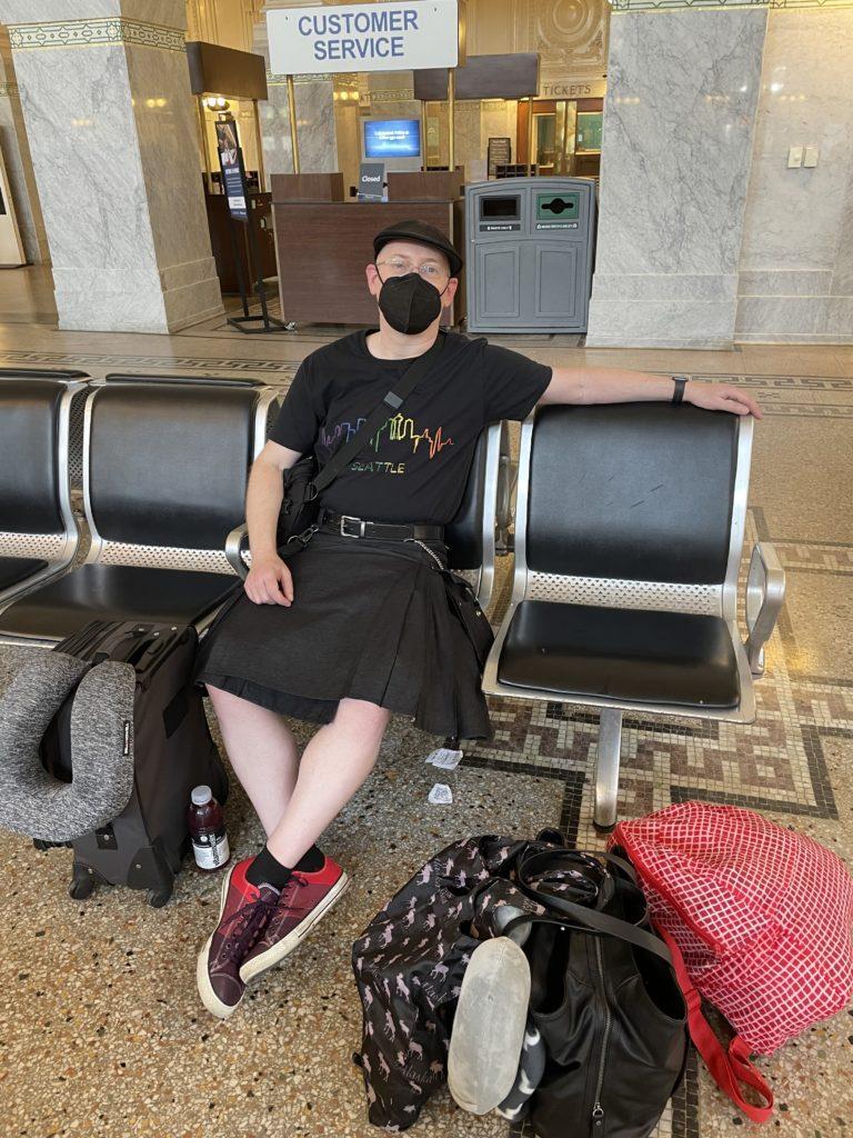 Me kicked back in a seat of the train station lobby. I’m masked and wearing a black cap, black t-shirt with rainbow Seattle logo, black kilt, and purple converse.