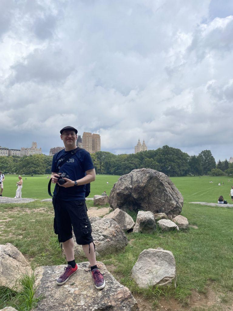 Me, wearing black shorts, purple Converse, and a navy blue shirt that says “RITOS”, holding a camera, and standing on a rock in front of a large grassy field in New York City’s Central Park. Some of NYC’s old buildings can be seen in the distance.