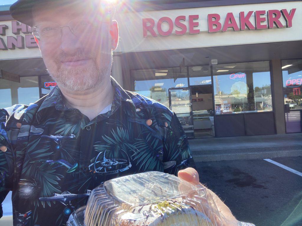 Me standing in a strip mall parking lot in front of Rose Bakery, holding a small container of cake.