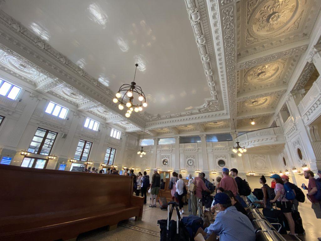 A wide angle view of King Street Station, showing the classic wooden benches, white and grey marble pillars, and ornate white ceiling panels. A line of passengers wait to board a train.