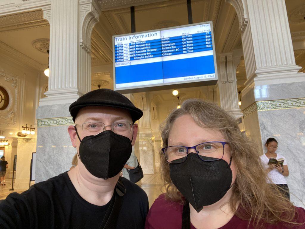 My wife and I, both masked, standing in Seattle’s King Street Station train station, all old white marble and ornate white ceilings. Behind and above us is an Amtrak departures board.