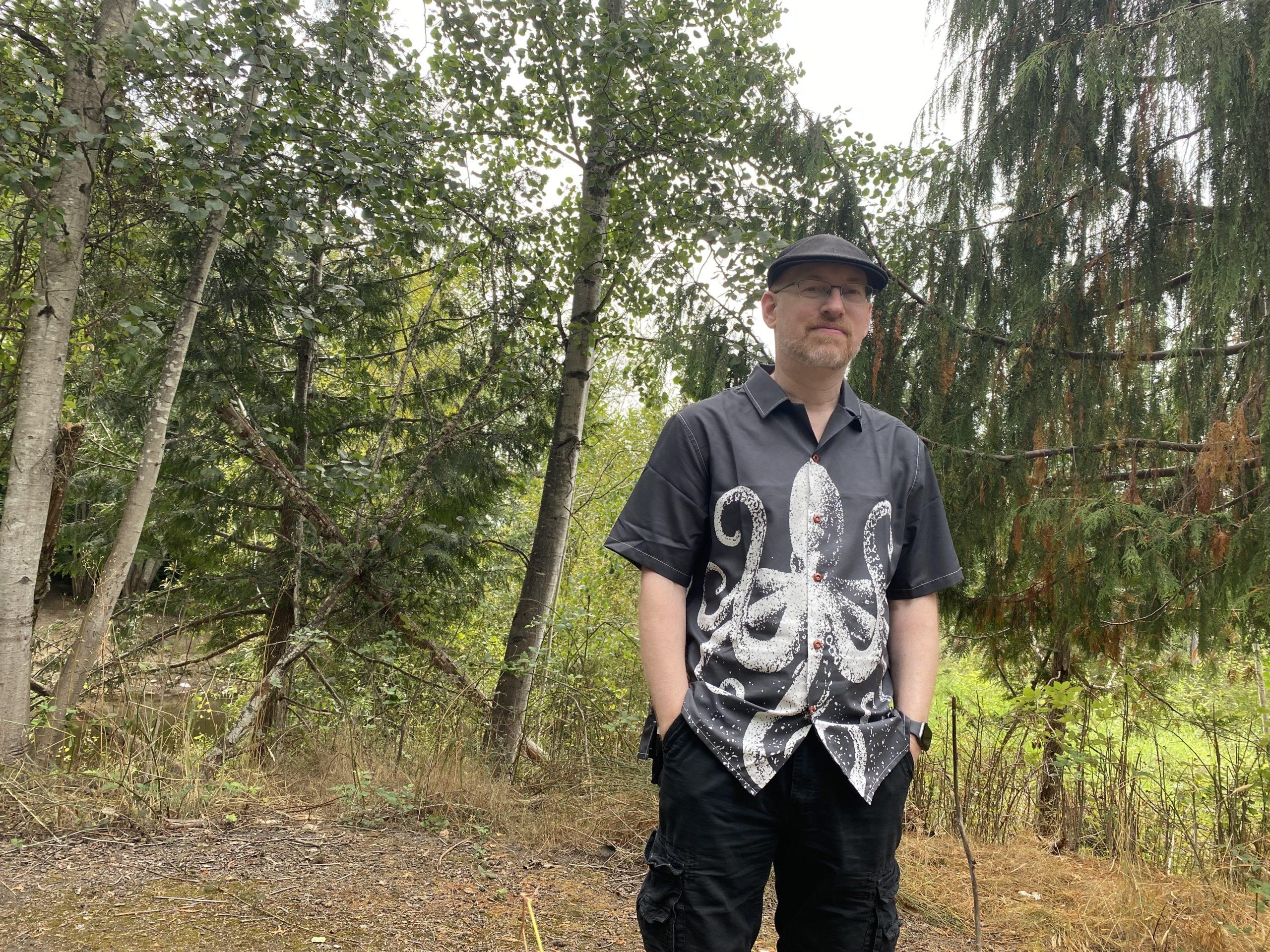 Me standing in a wooded area, wearing black shorts, a black cap, and a black button-up short-sleeve shirt with a black-and-white print of an octopus.