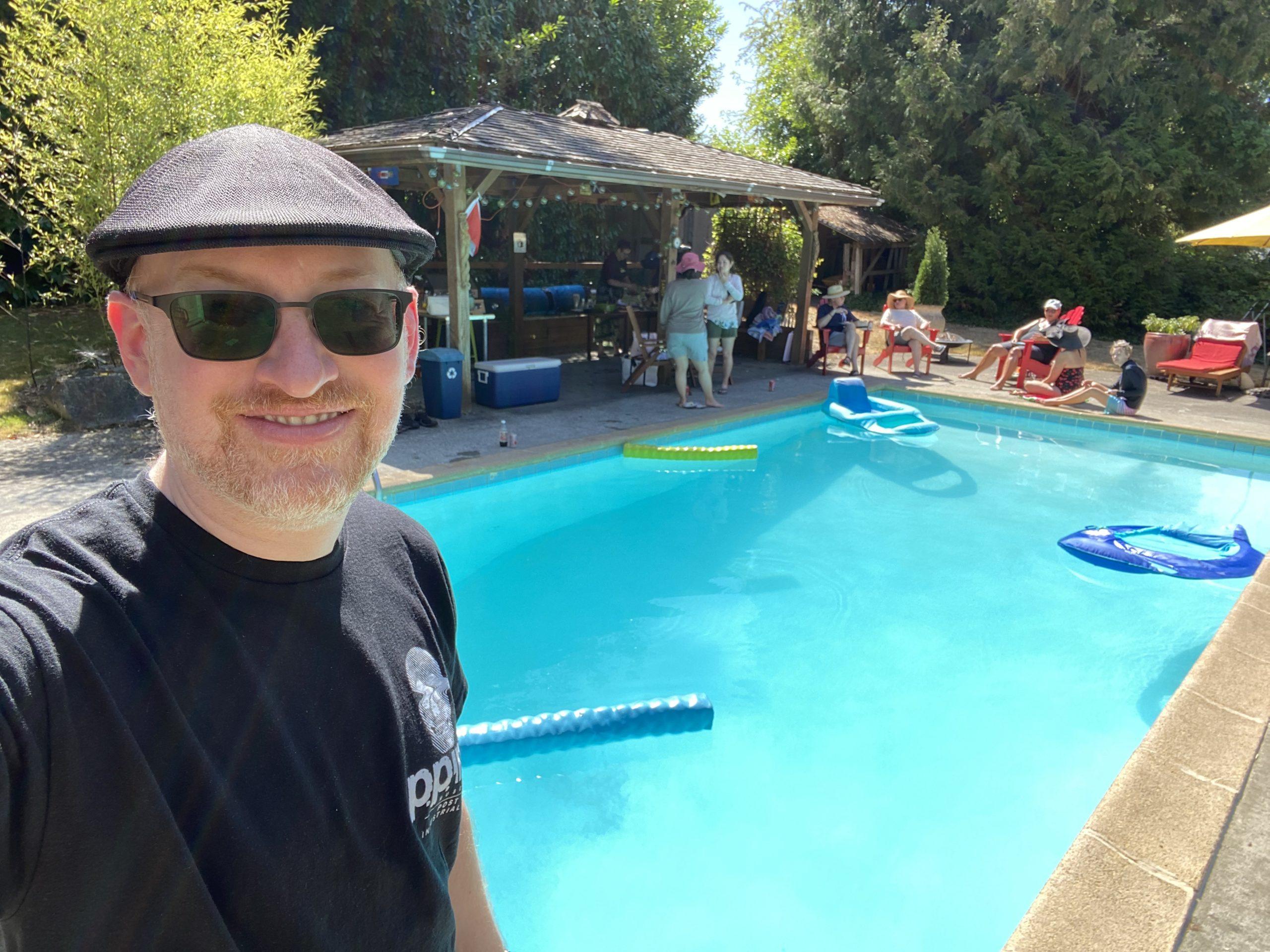 Me standing in front of an in-ground pool on a sunny day. On the far side of the pool is a sheltered area decorated for a party. People can be seen chatting, standing or sitting near the pool, some in the sun, some in the shade.