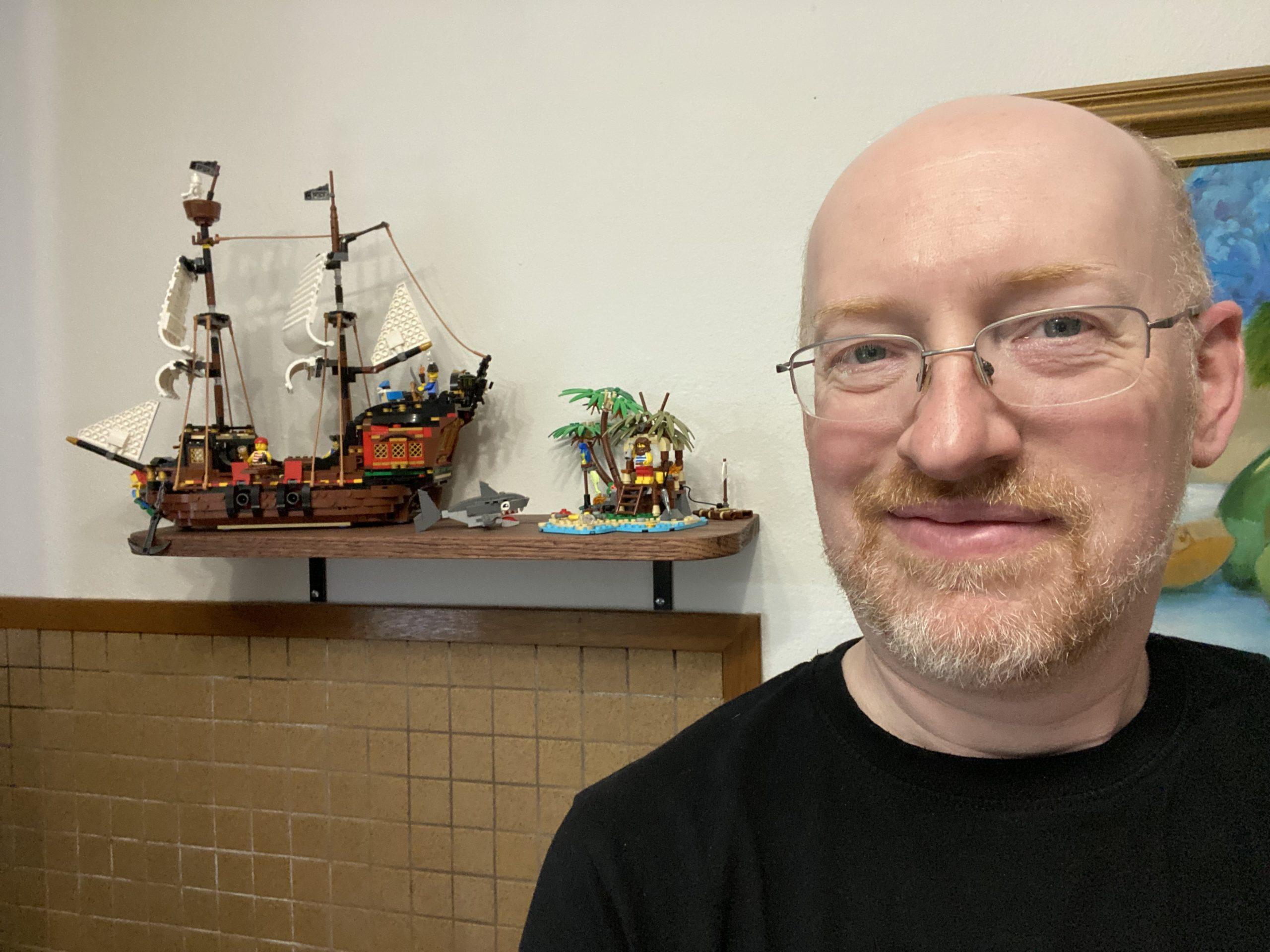 Me standing in front of a wall; on a shelf mounted on the wall is a Lego pirate ship, shark, and small desert island with a castaway.