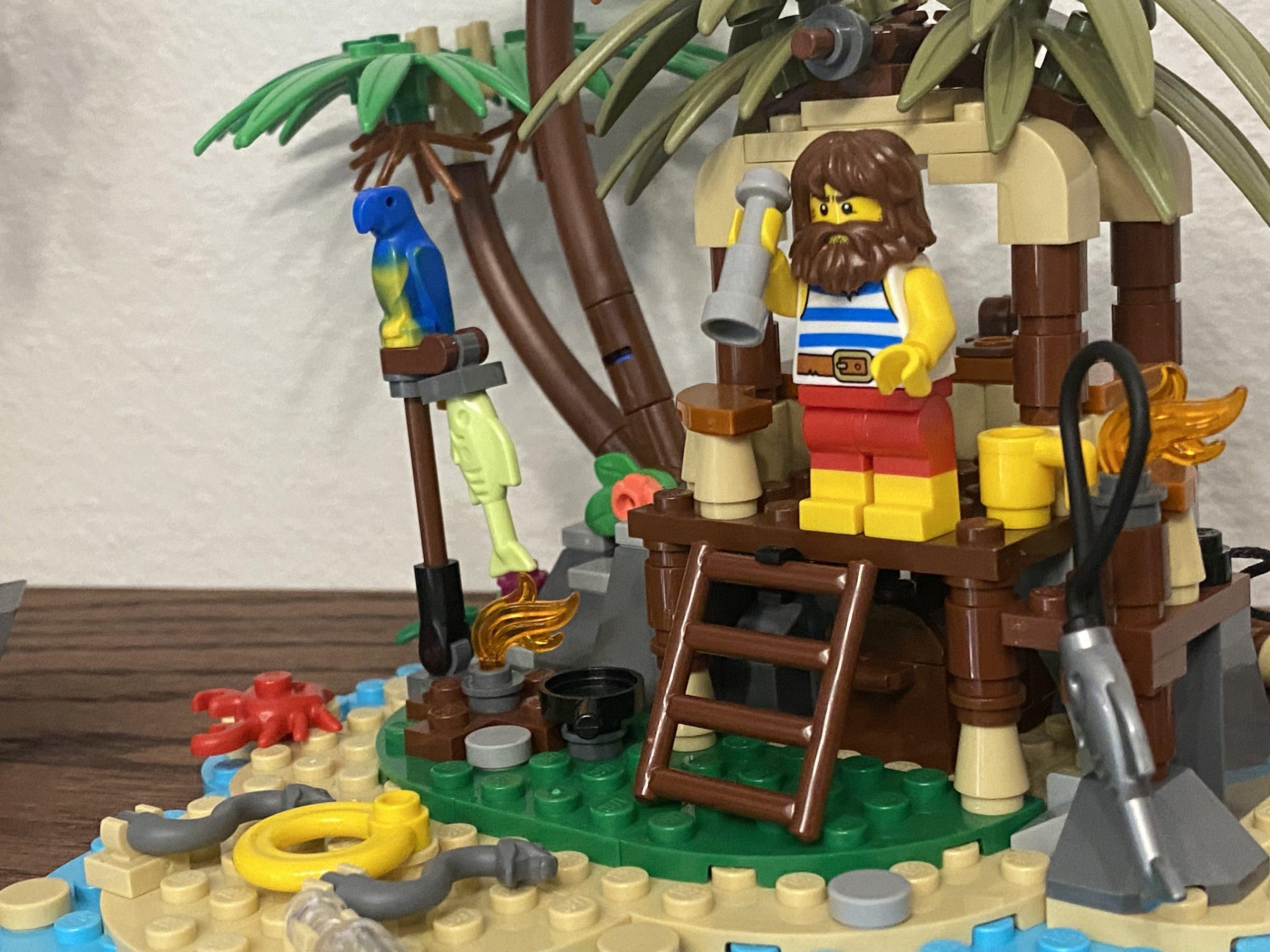 A Lego island with a castaway standing on a platform under trees. There is a parrot sitting on a perch, a fishing pole with a fish, and snakes on the beach by the water.