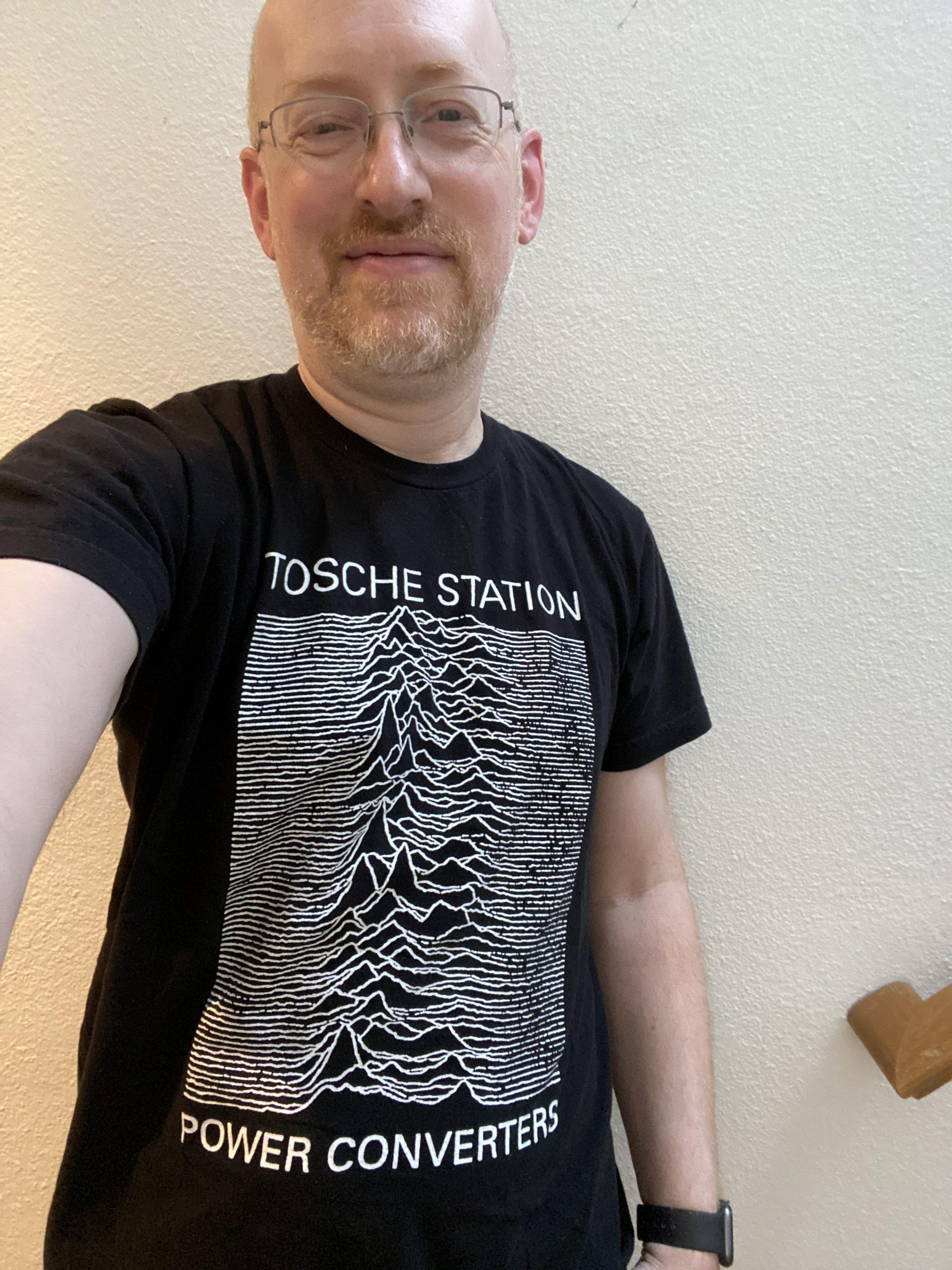 Me standing in front of a white wall, wearing a black t-shirt riffing on the classic Joy Division Unknown Pleasures album artwork, only with text that says Tosche Station / Power Converters.
