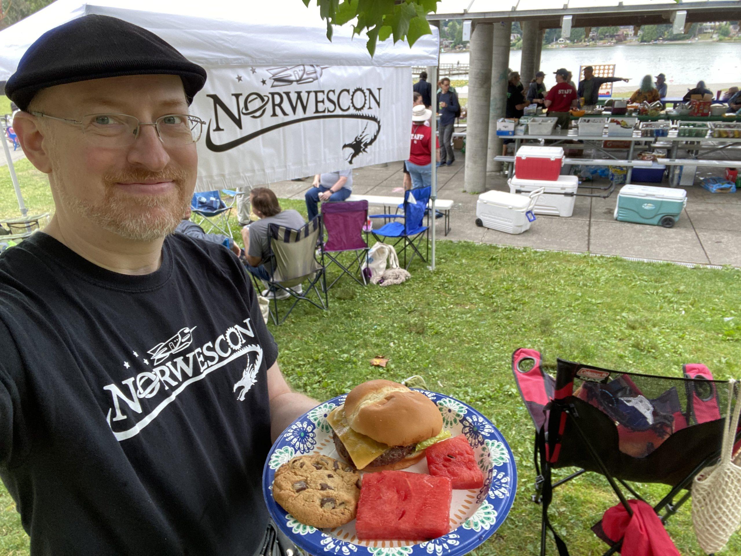Me standing in a park near a picnic shelter with people and supplies at the tables. Behind me is a pop-up canopy with a Norwescon logo banner hanging from it. I'm wearing a Norwescon shirt, and holding a plate of food.