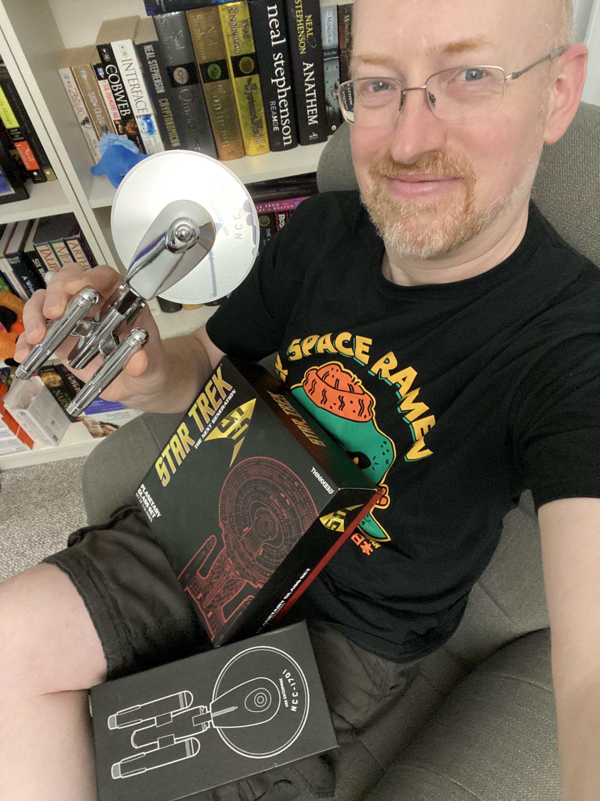 Me sitting in a chair in front of a bookcase, with boxes for Star Trek glassware and a pizza cutter in the shape of the original Enterprise in my lap, holding the pizza cutter.