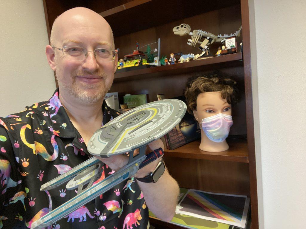 Me standing in front of a bookcase. Visible on the bookcase is a mannequin head with curly brown hair, wearing a pastel rainbow face mask. I'm holding a model of the USS Cerritos from Star Trek Lower Decks.
