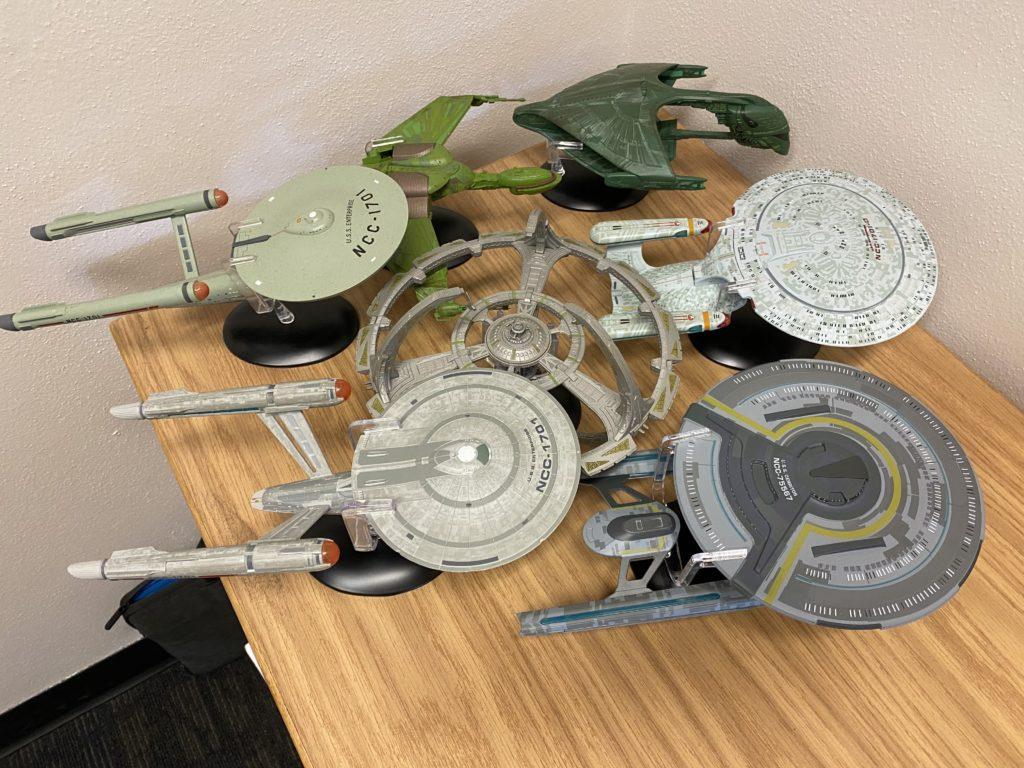 A collection of Star Trek ship models on my work desk. Visible is the original TOS USS Enterprise, the TOS Klingon Bird of Prey, the TNG Romulan Warbird, the TNG 1701-D Enterprise, Deep Space Nine, the Strange New Worlds USS Enterprise, and the Lower Decks USS Cerritos.