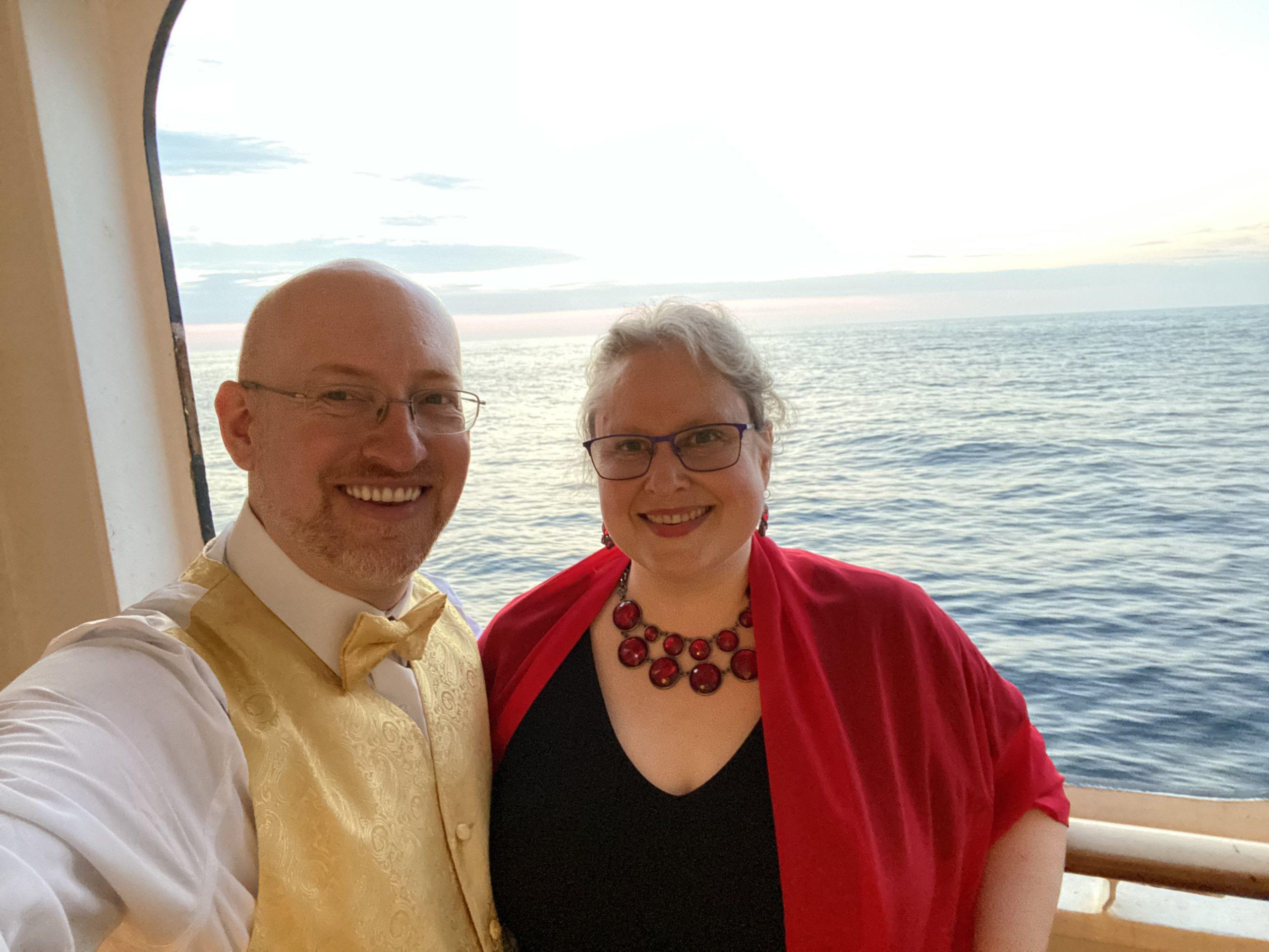 My wife and I in formal dress on the balcony of our stateroom with a calm ocean behind us. I'm wearing a white dress shirt with gold vest and bow tie, she's wearing a black dress with red wrap and jewelry.