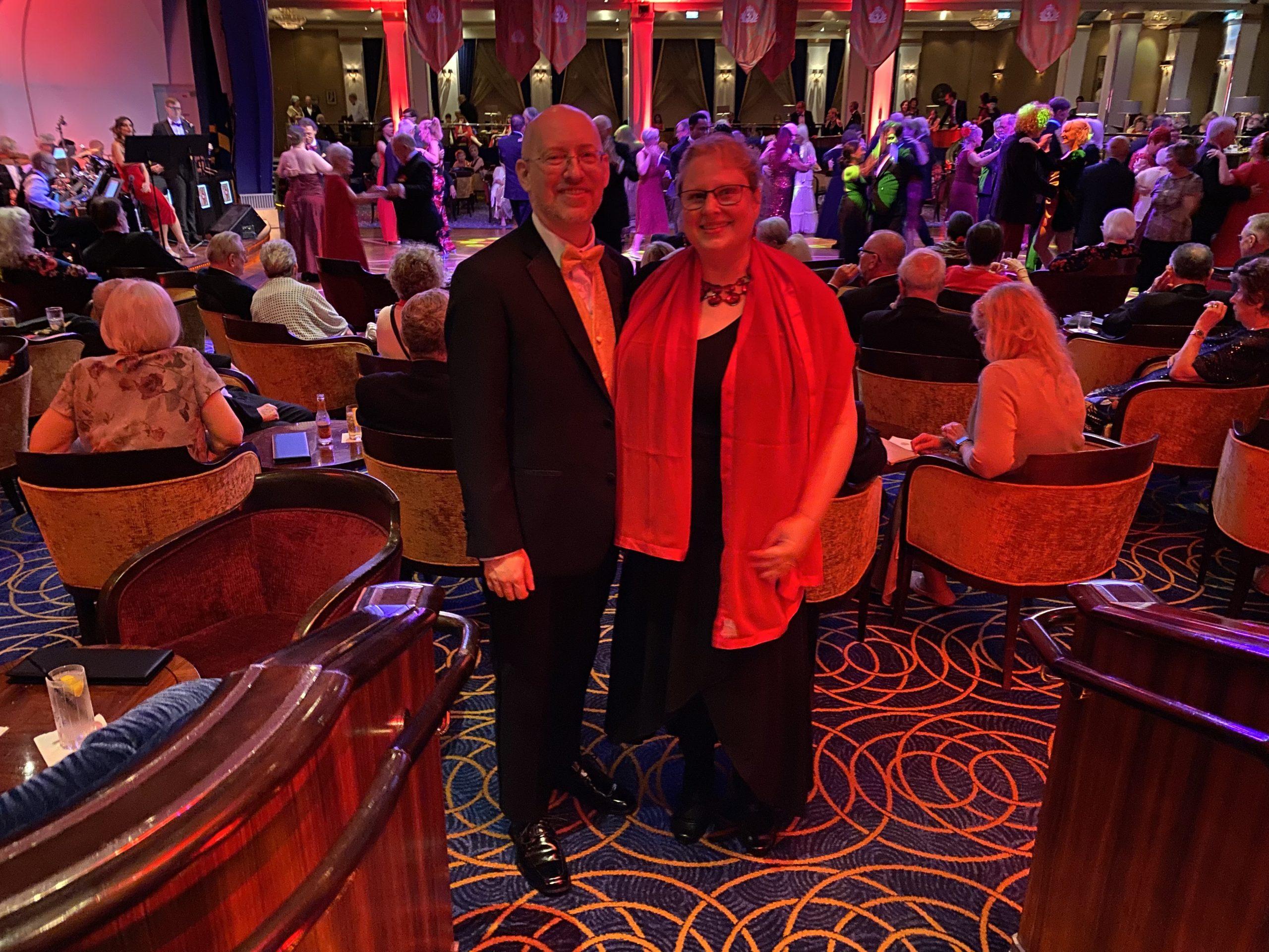 My wife and I in formal dress at a fancy event. I'm wearing a tux with gold vest and bow tie, she's wearing a black dress with red wrap and jewelry. Visible behind us are a number of people ballroom dancing to a live band while other people sit in chairs and watch.