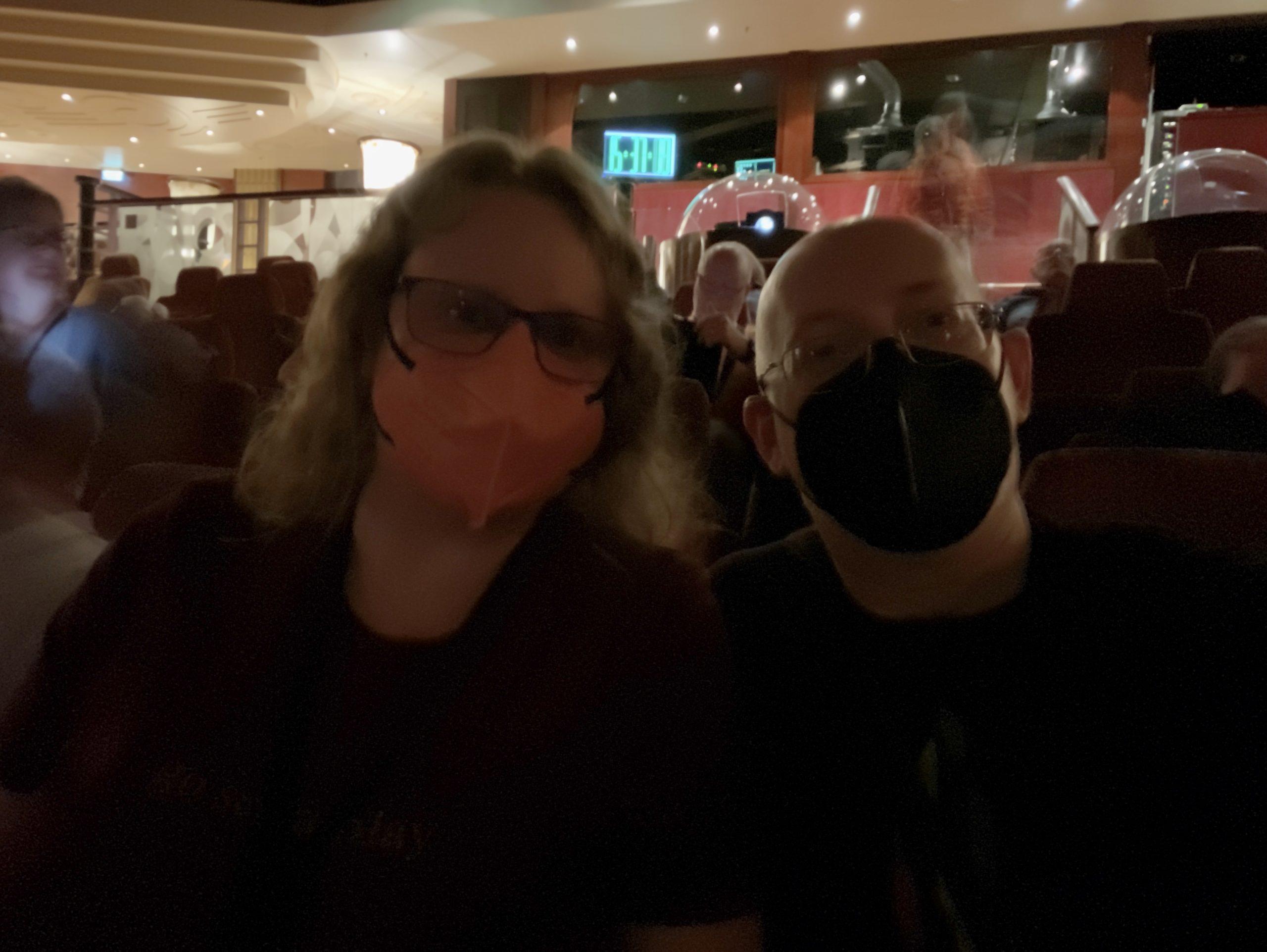 A dark photo of my wife and I, both wearing masks, sitting in auditorium seats. People around us are half-visible and blurred due to the long exposure.