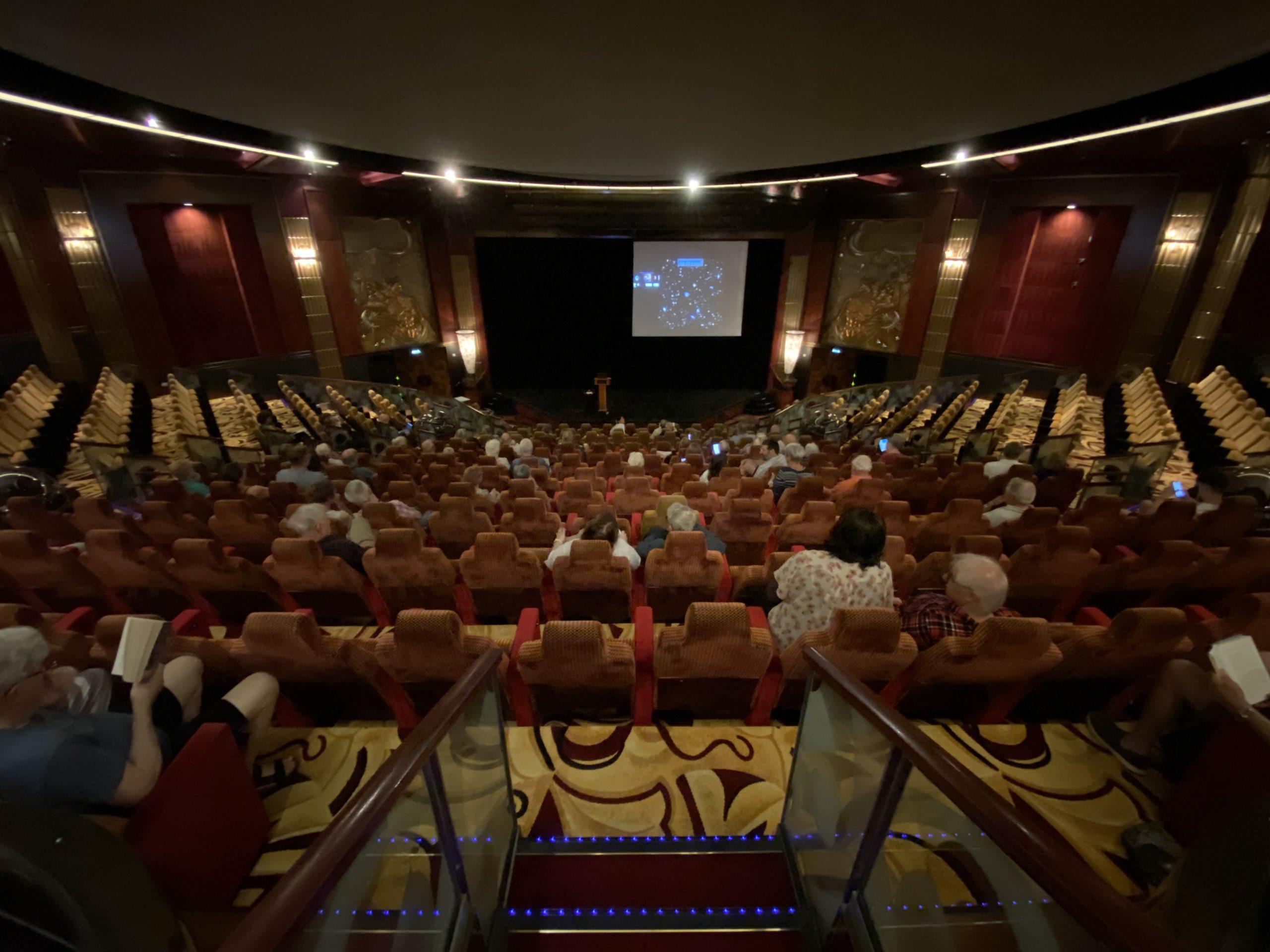 A view of the lecture hall and planetarium from the back, with people starting to take seats and an image of a starfield on the screen on the stage.