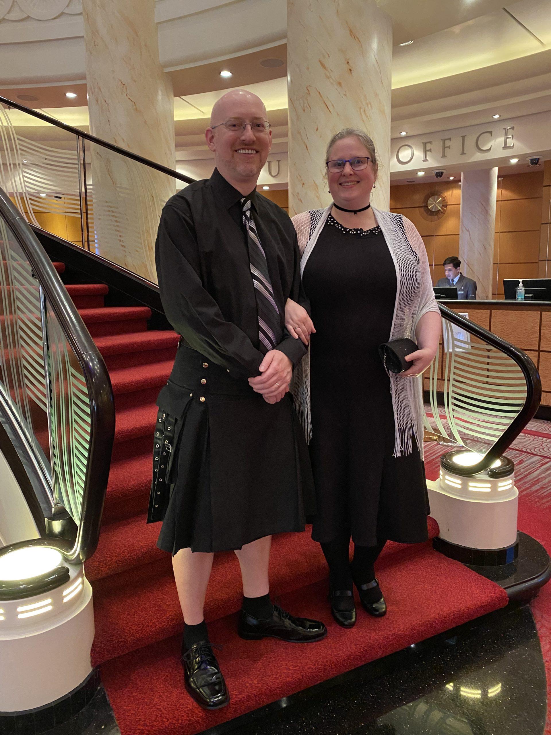 My wife and I standing at the bottom of the ship's grand stairway. I'm wearing a black long-sleeve shirt, black and grey necktie, and black kilt; she's wearing a black dress with silvery-grey wrap and a black choker necklace.