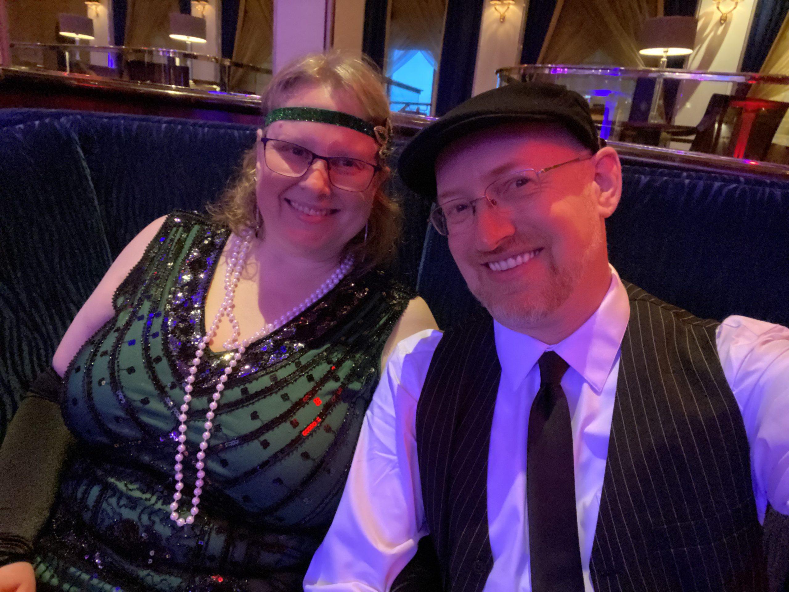 My wife and I in 1920s-style outfits. I'm wearing a white shirt with black tie, pinstriped black vest, and black cap, she's wearing a green flapper dress with black sequins, a long string of pearls, and a green sequined headband.