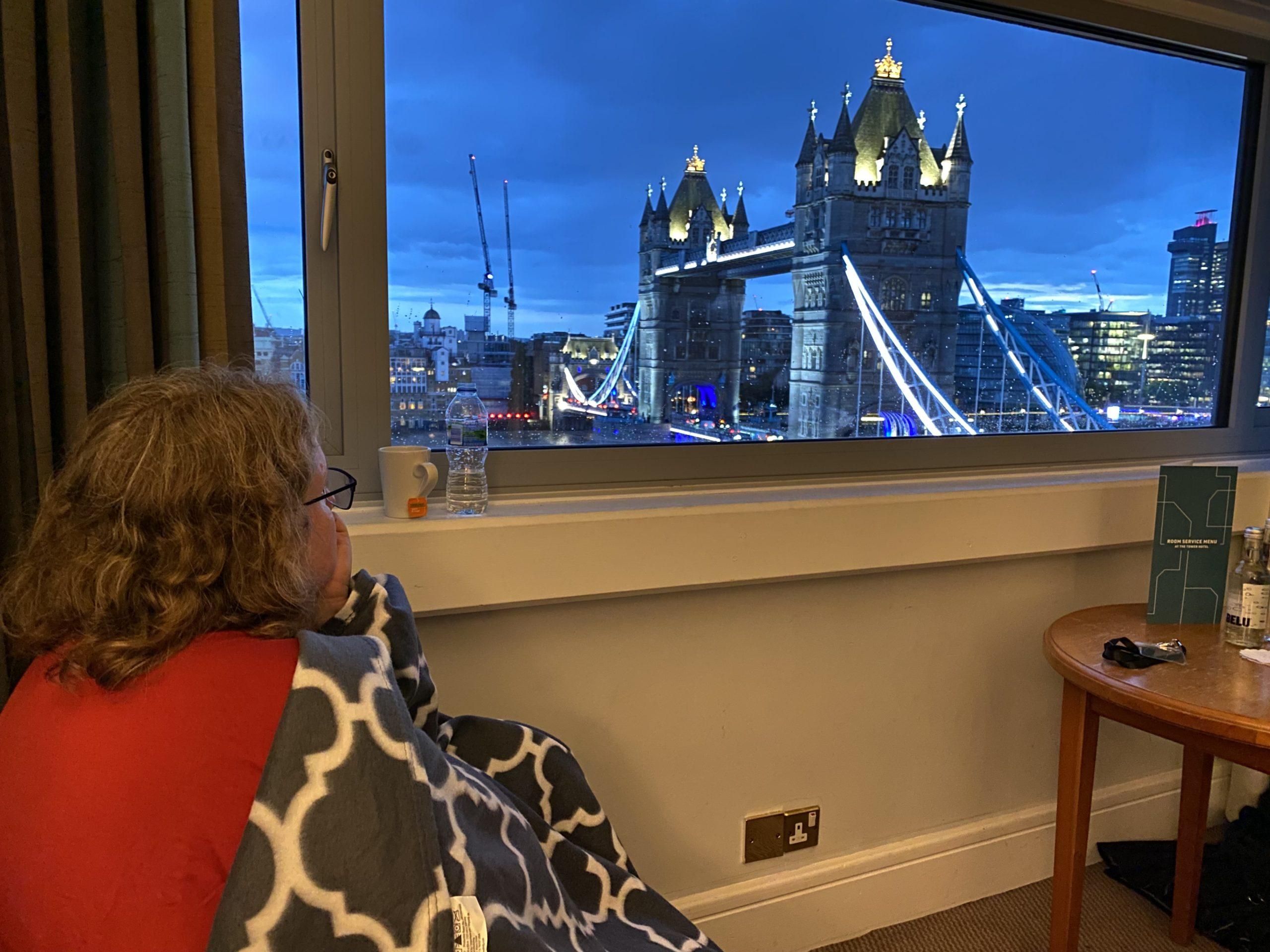 My wife curled up in a chair under a blanket, looking out the hotel winow at the Tower Bridge lit up as the sky darkens into twilight.