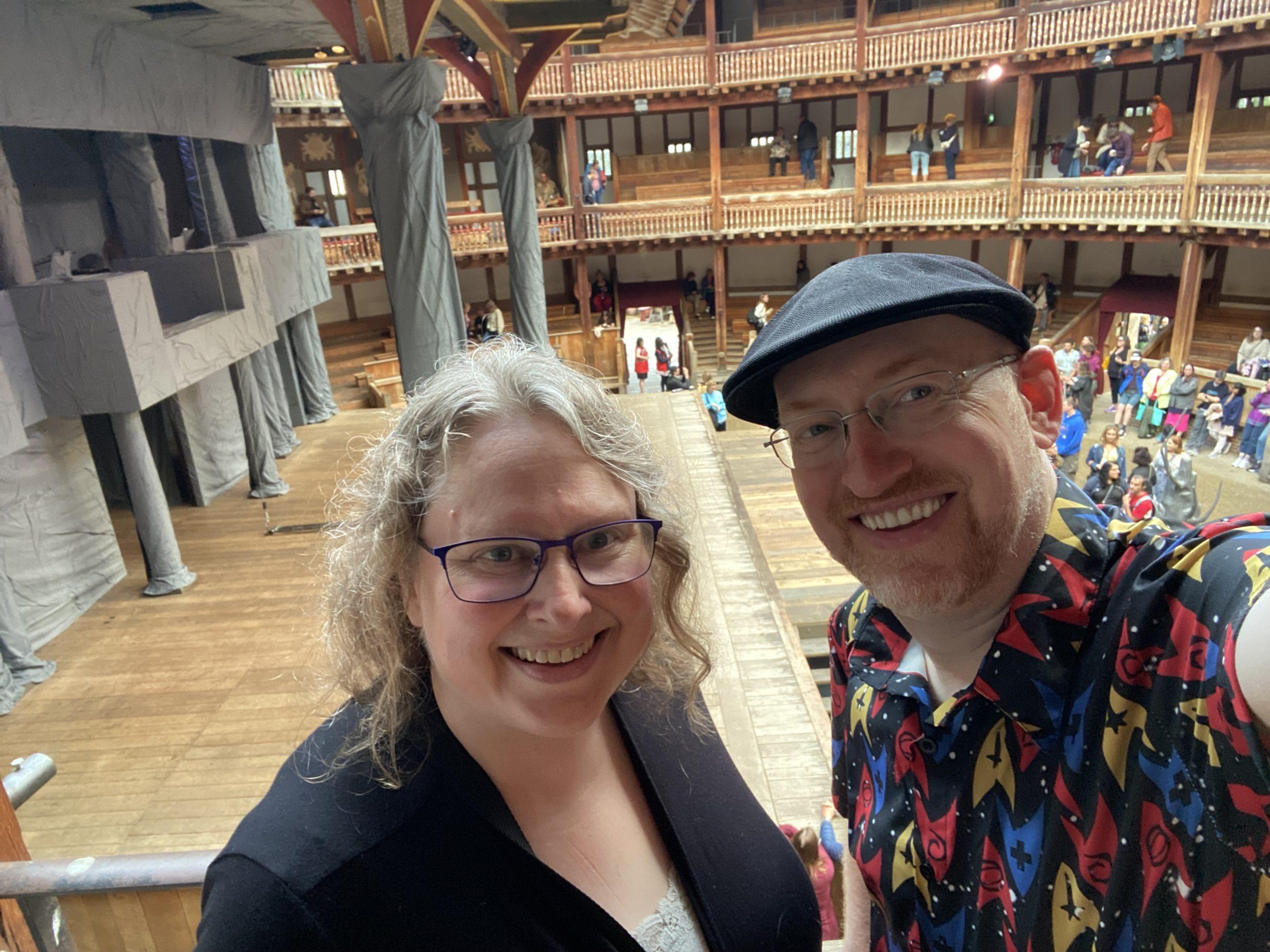 My wife and I at Shakespeare's Globe Theater in London. We're in one of the upper galleries overlooking the stage and floor area. Other audience members are finding their spots for the performance behind us.