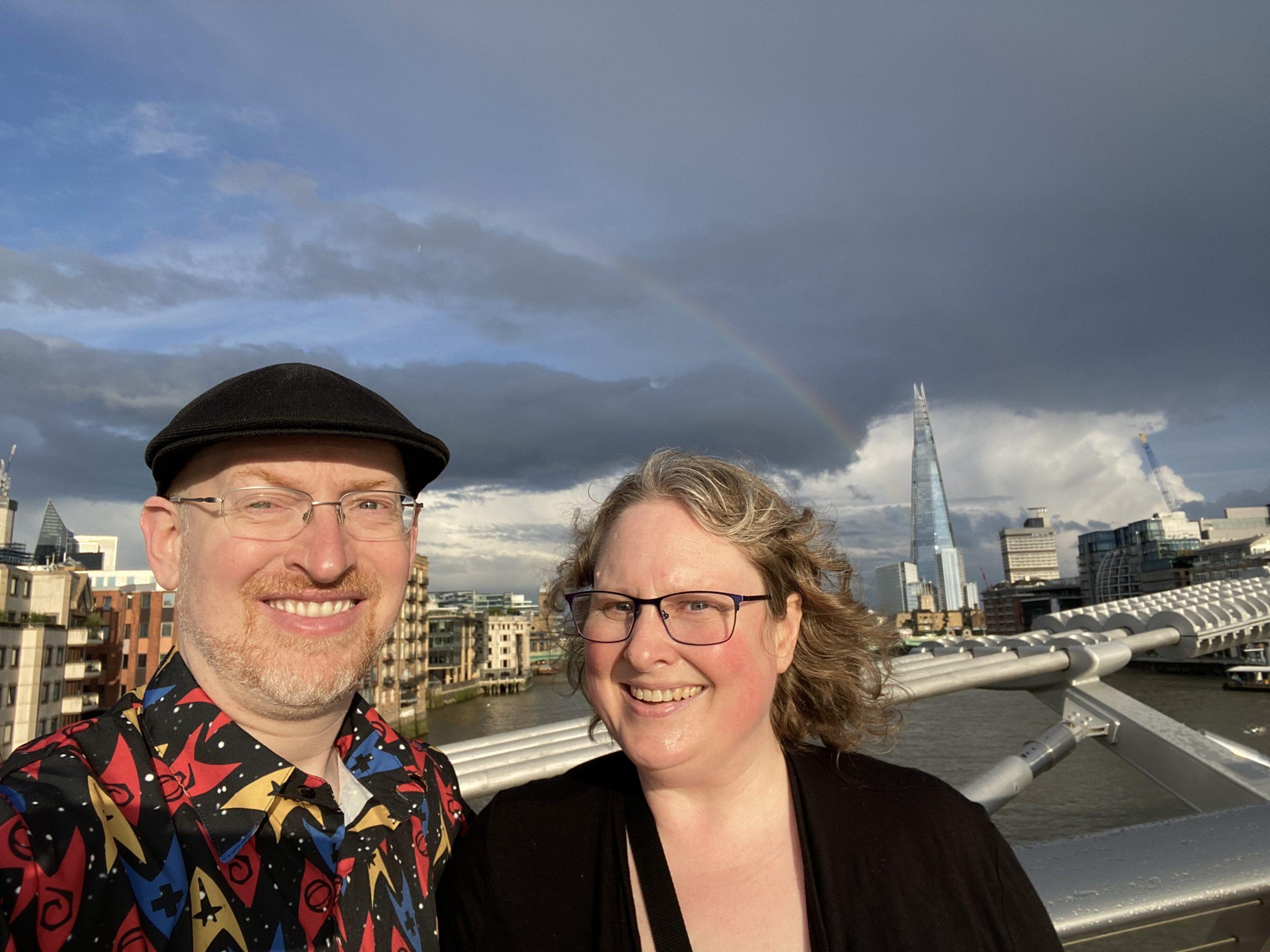 My wife and I, both kind of squinty, on the Millennium Bridge with a rainbow arcing over London visible behind us.