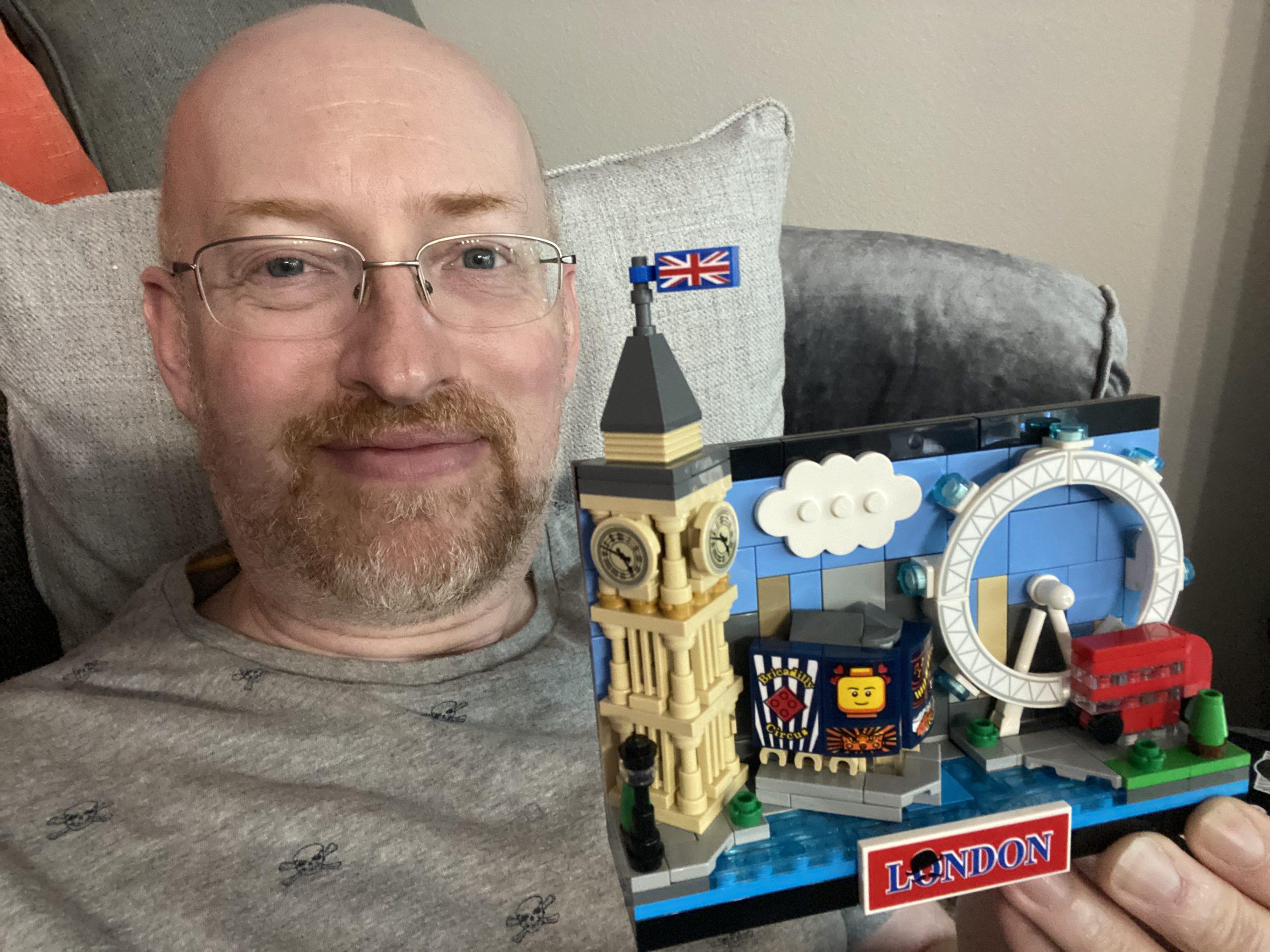 Me on the couch holding a small Lego model of London featuring Big Ben, Picadilly Circus, the London Eye, and a classic red double-decker bus.