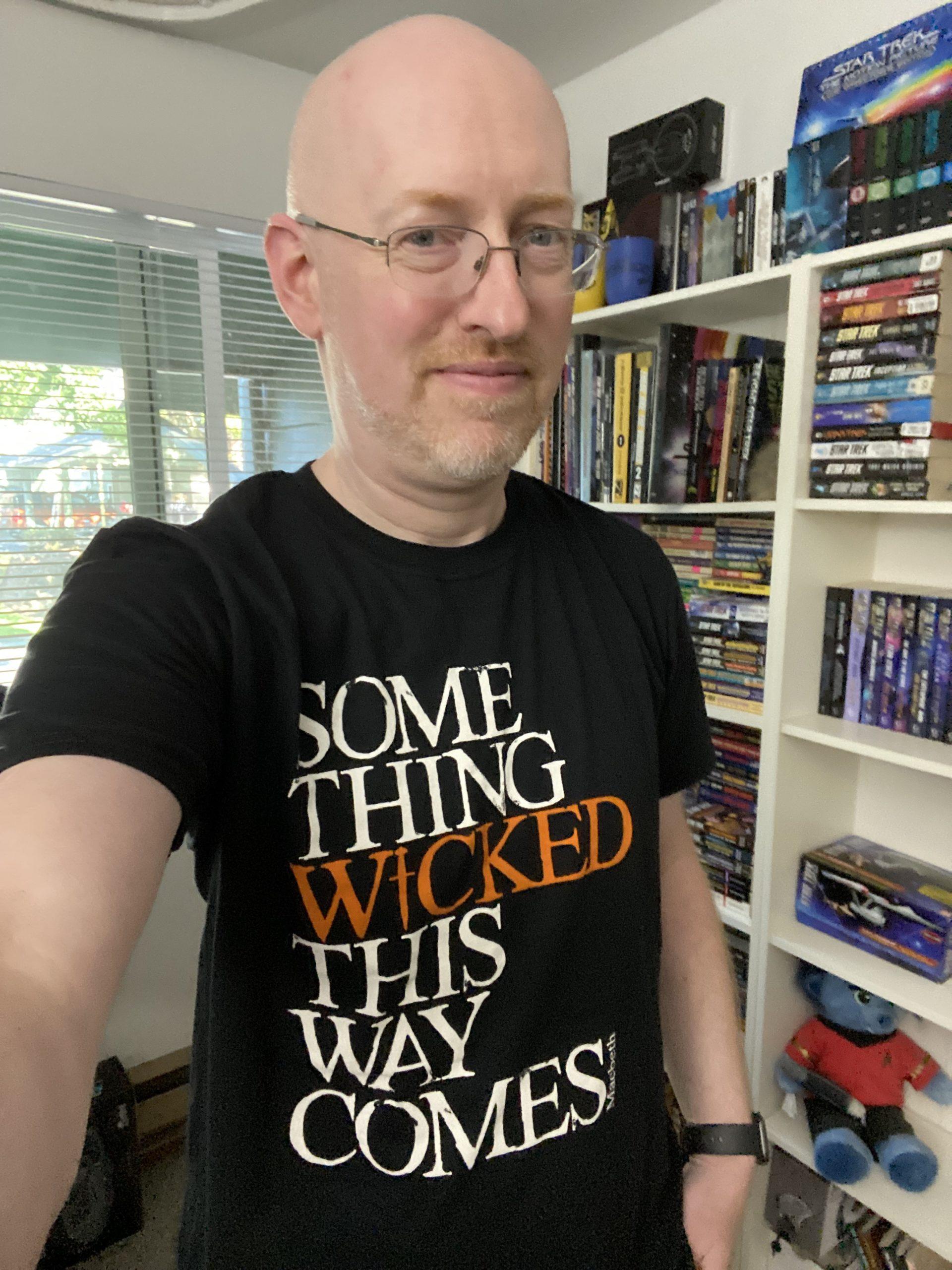 Me wearing a black t-shirt with slightly distressed white text that says 'something wicked this way comes'; the word 'wicked' is printed in orange.