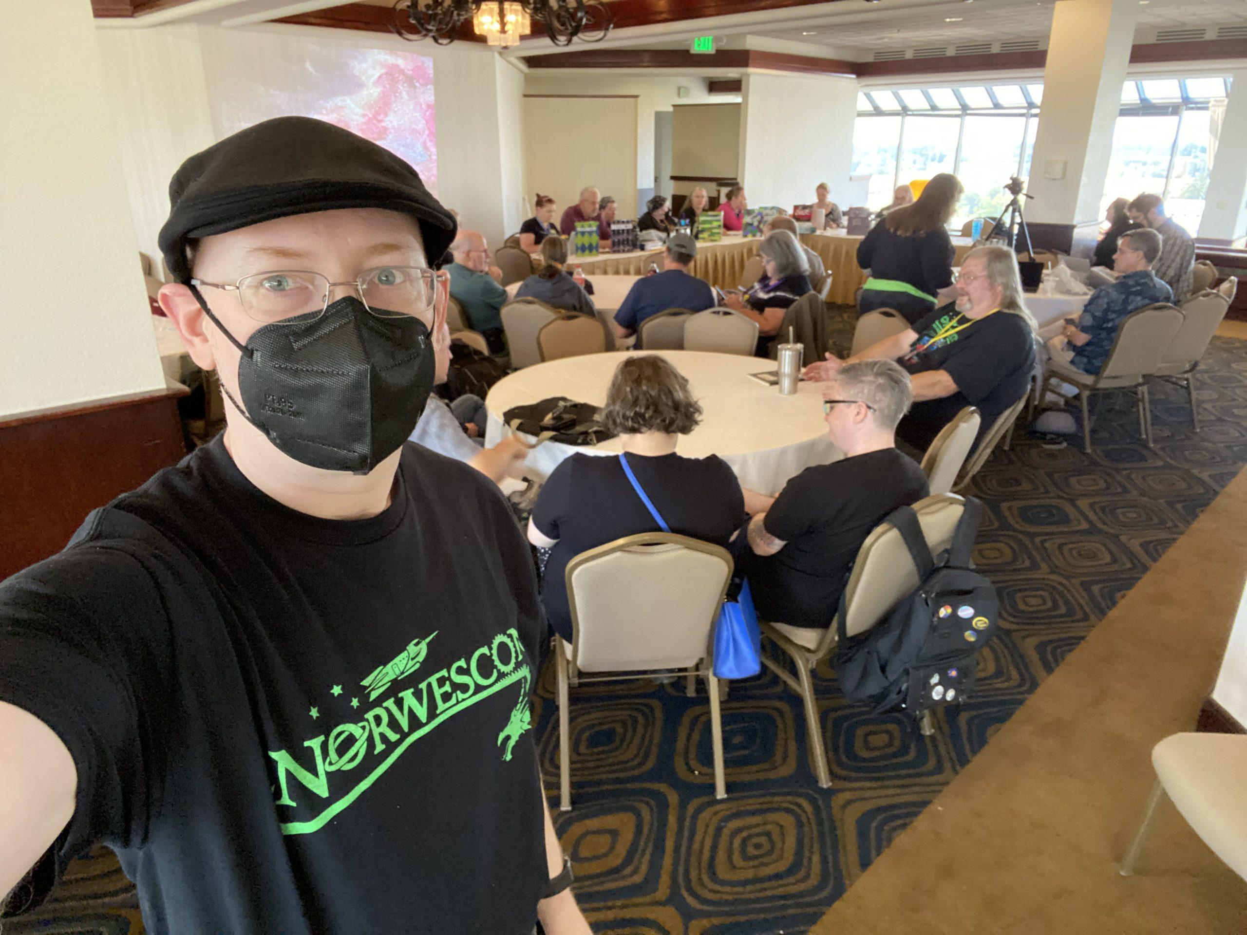 Me, wearing a black cap, mask, and t-shirt with the Norwescon logo in green, in a hotel conference room with other people sitting around a number of round tables.