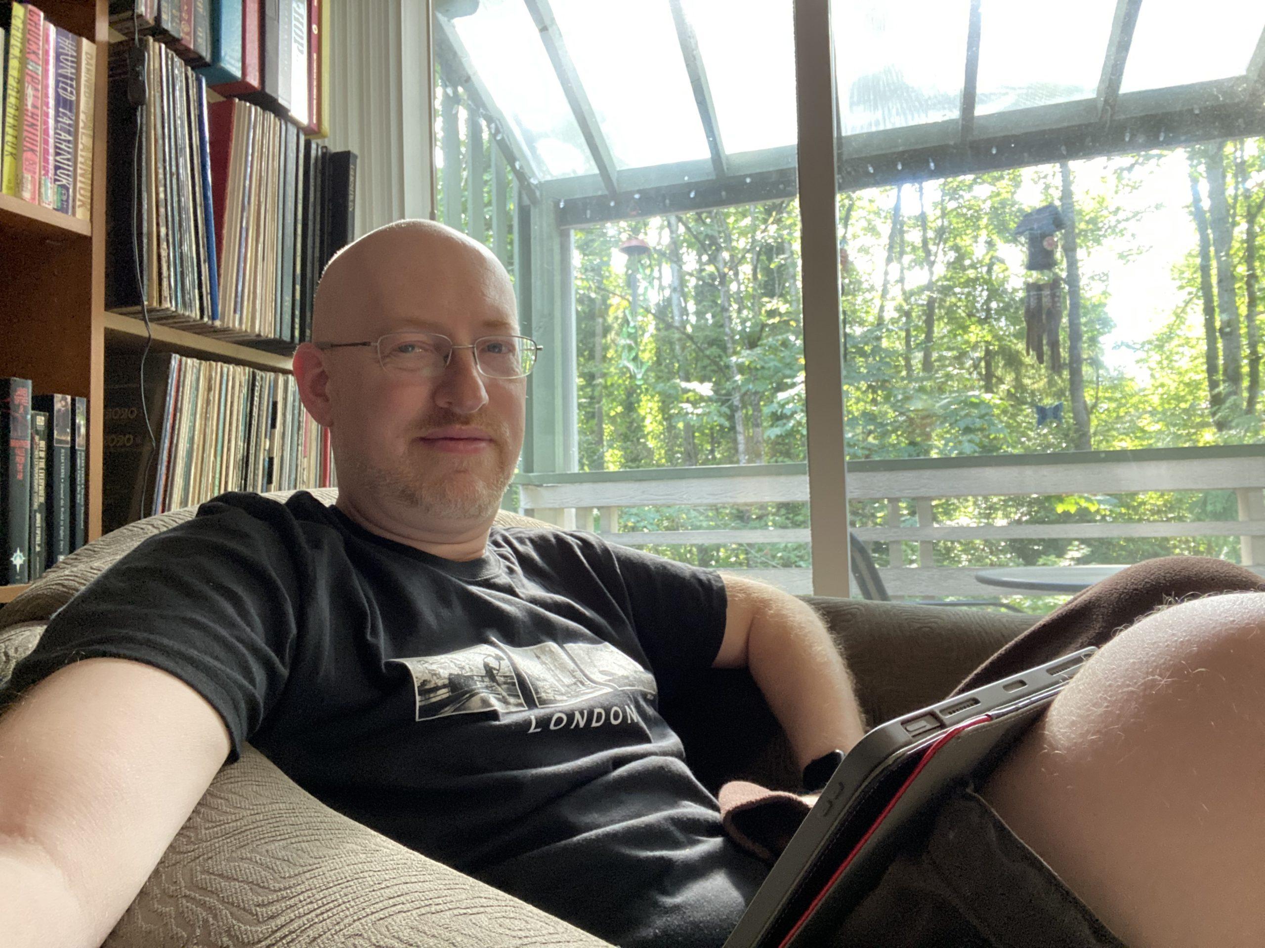 Me sitting in a comfortable chair, with my iPad on my lap, in front of a window looking out on sunlit green trees.