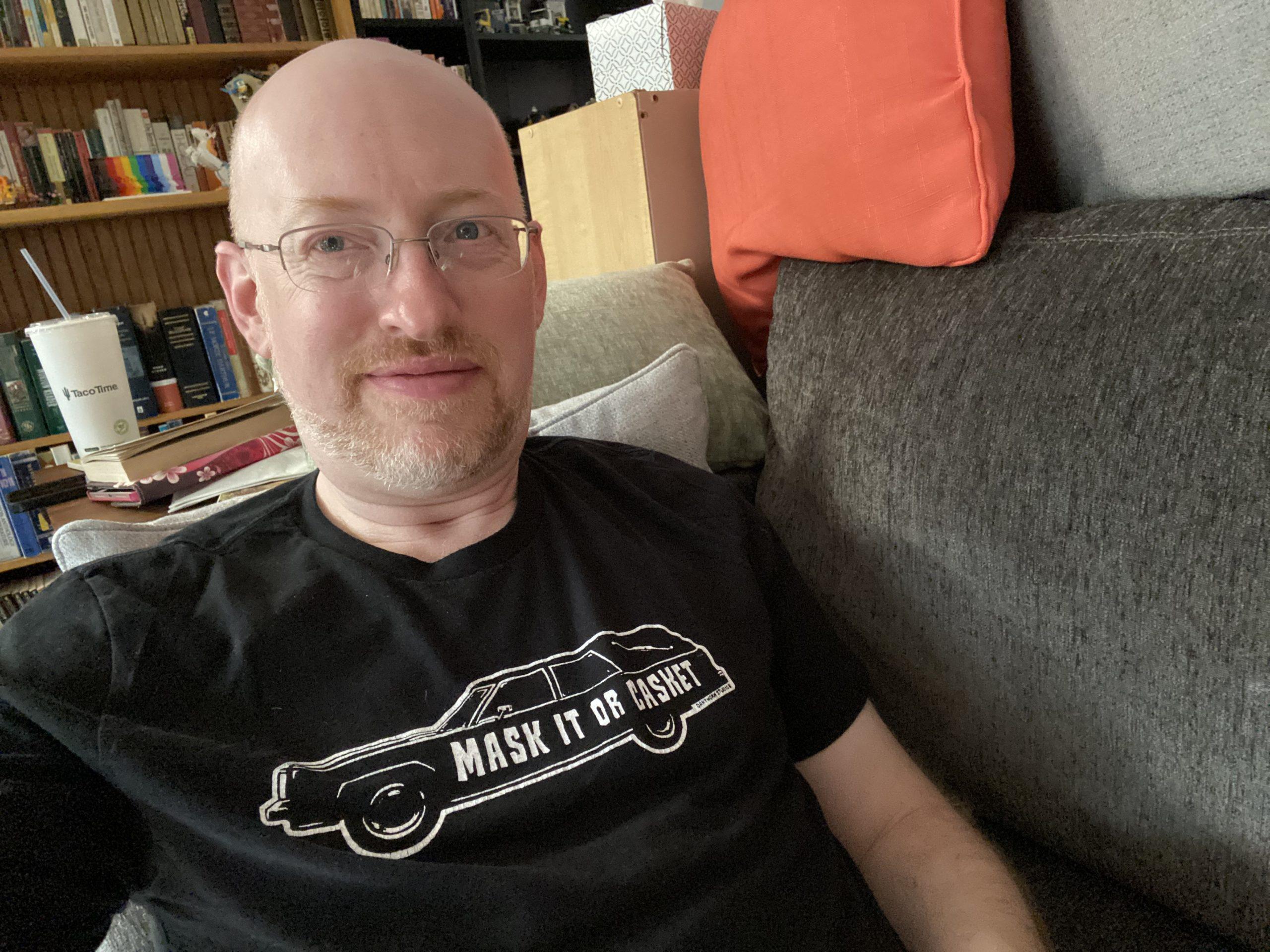 Me sitting on our couch, wearing a black t-shirt with a graphic of a hearse with the words 'mask it or casket' written across the side.