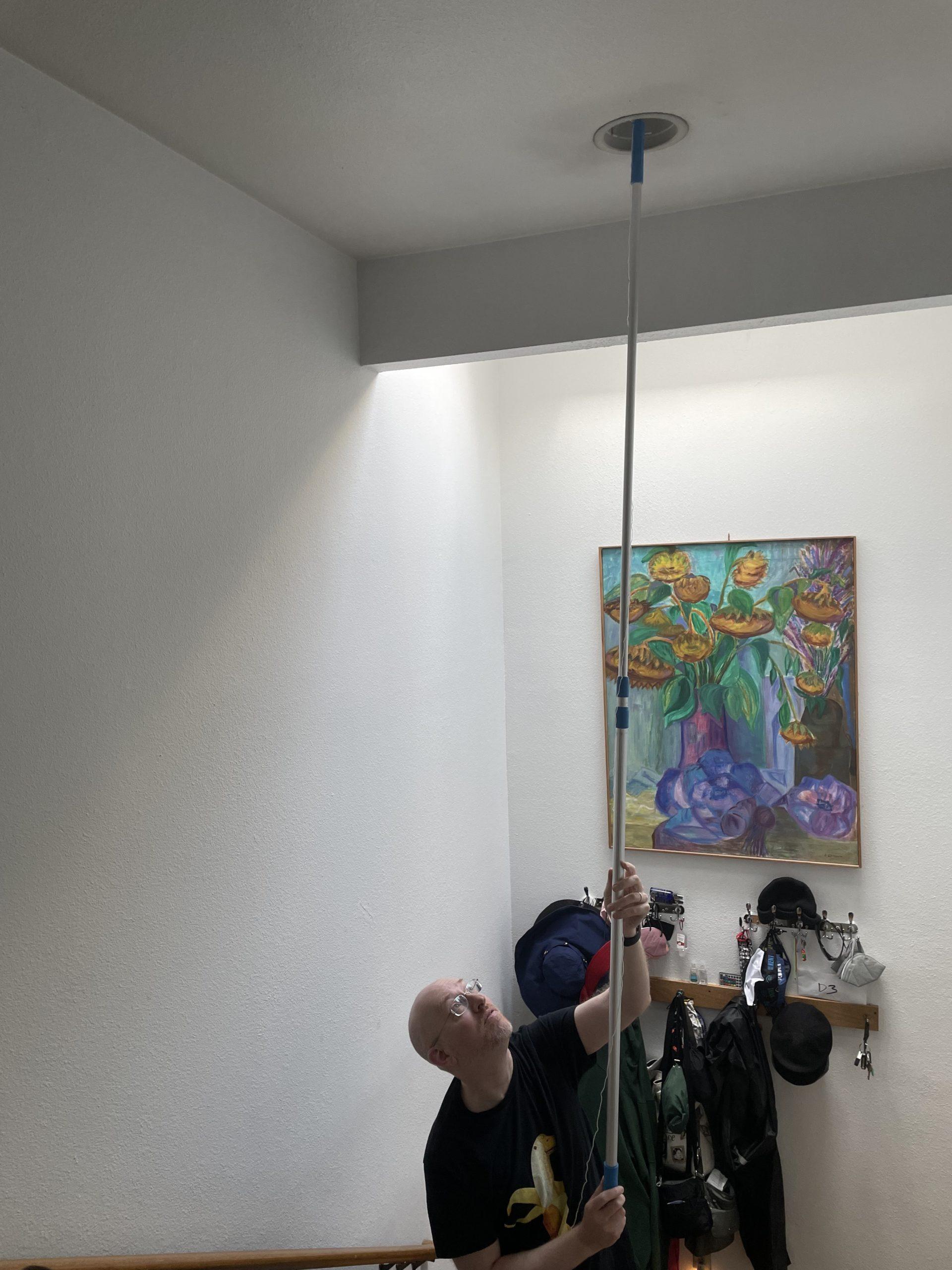 Me standing in an entryway with a high ceiling, using an extension pole to replace a ceiling-mounted lightbulb.