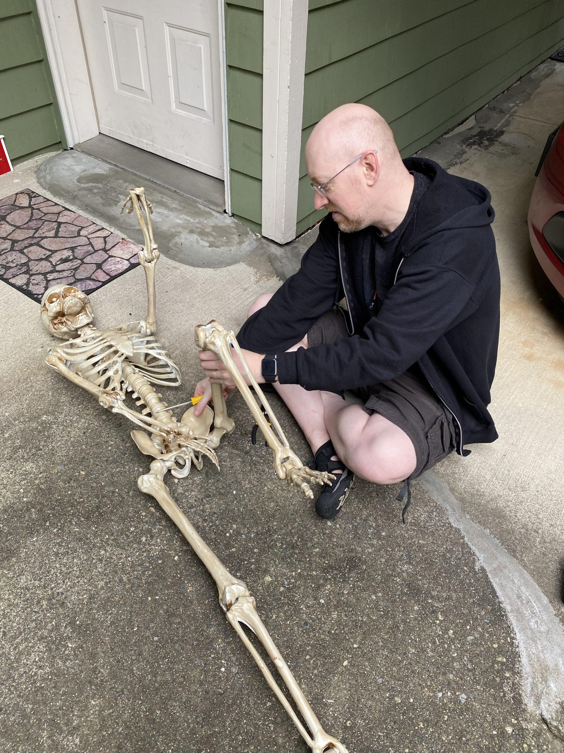 Me sitting on the ground in front of our front door, adjusting a poseable decorative skeleton on the ground in front of me.