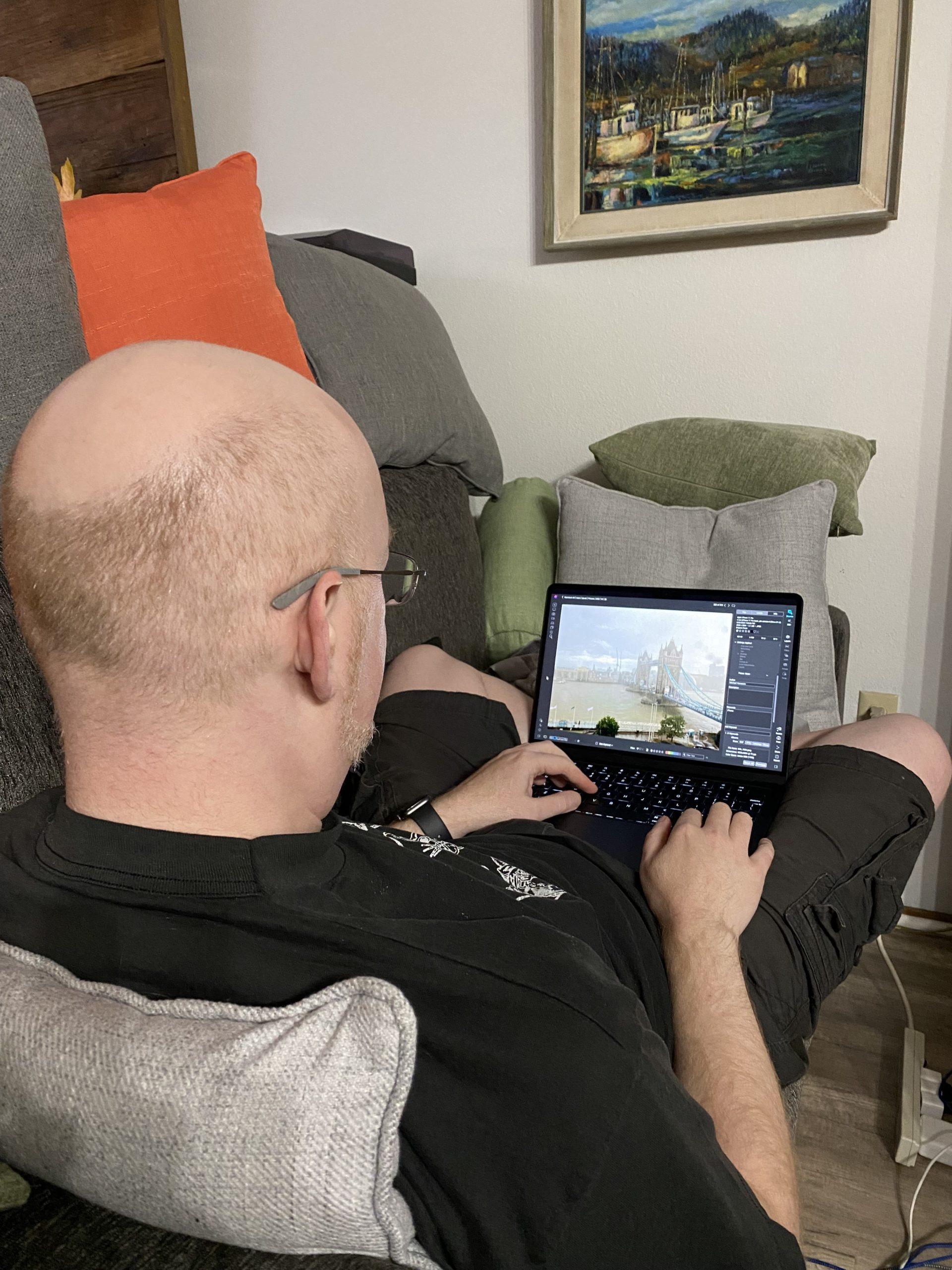 Me from behind, sitting on our couch with my comptuer in my lap, with a photo of London's Tower Bridge visible on the screen.