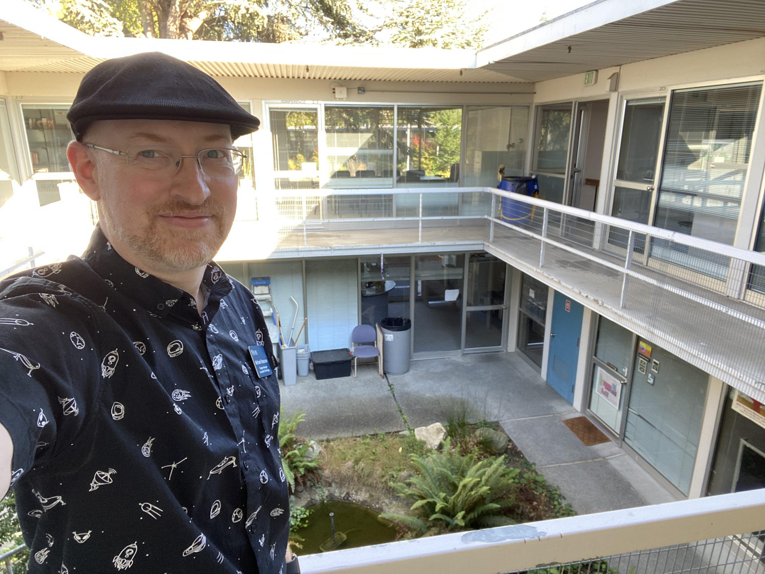 Me standing on a second-floor walkway in a small square building with a courtyard in the center. In the courtyard is a small pond surrounded by plants. The sun is shining through the open roof.
