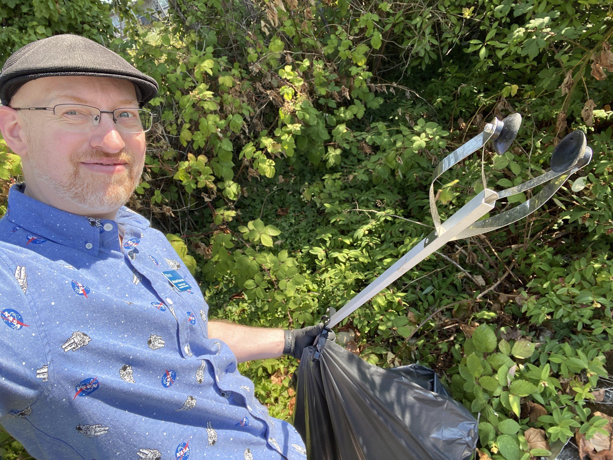 Me standing in front of some bushes and ivy, holding a full trashbag and a 'grabber' used for picking up trash.
