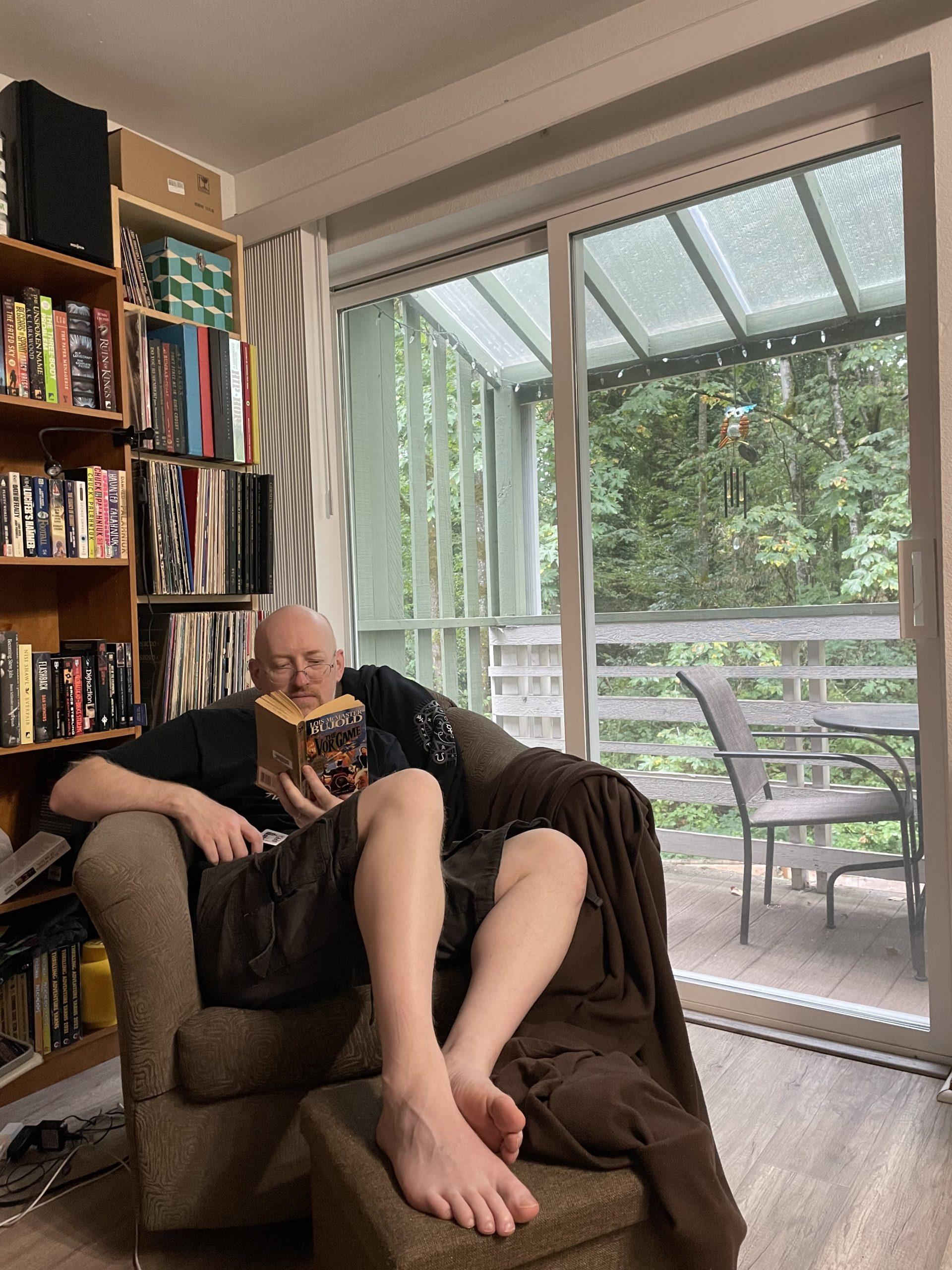 Me sitting in our living room in a comfortable chair with my feet on an ottoman, reading a book. Books and vinyl records are visible on the shelves behind me, and green trees can be seen through a glass sliding door leading onto a deck.