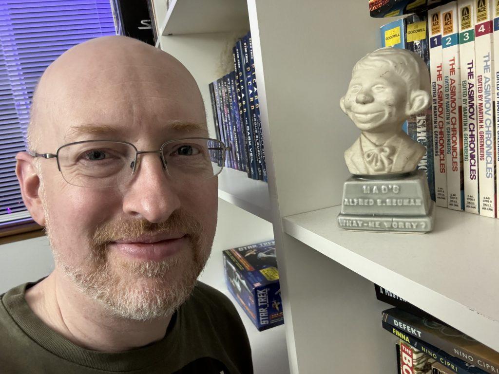 Me next to a white bookshelf with a number of science-fiction books visible. Next to my head is a small bust of Mad Magazine’s Alfred E Neuman with his ‘what, me worry?’ phrase on the pedestal.