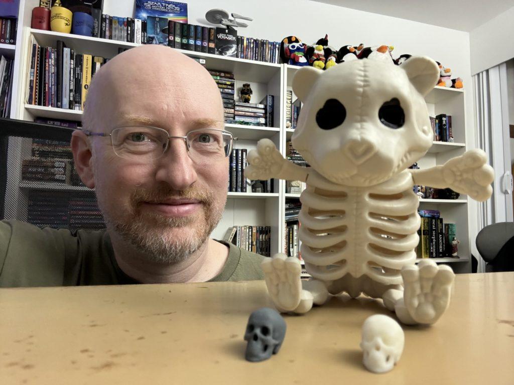 Me crouched in front of my desk. In the foreground, on the desktop, is a plastic Halloween teddy bear skeleton, with two tiny plastic human skulls at its feet.