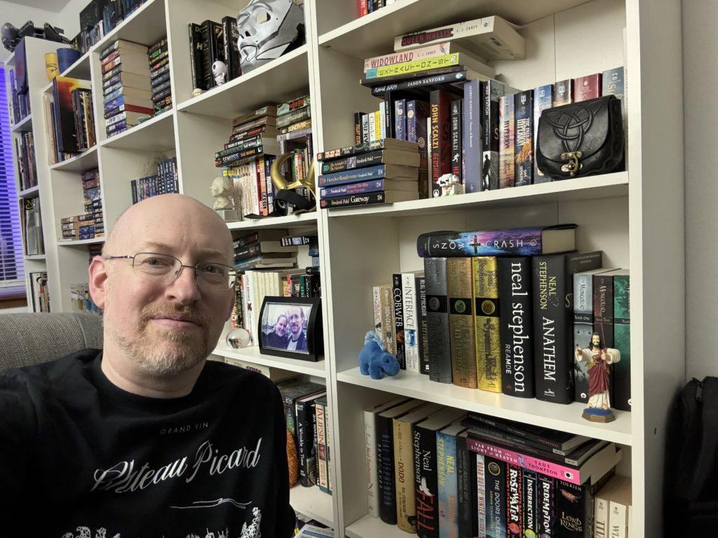 Me in front of four identical white IKEA bookshelves. Every shelf has books laid sideways atop the books that are shelved properly.