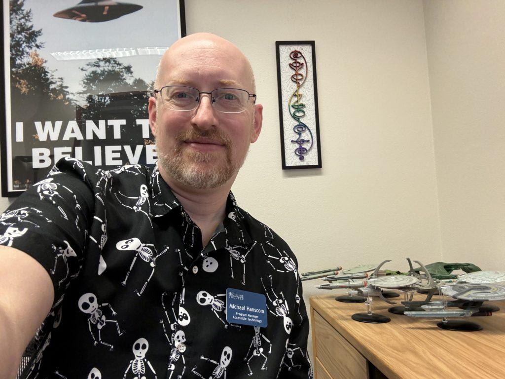 Me in my office at work, wearing a black short-sleeve button-up shirt with cartoonish dancing skeletons.